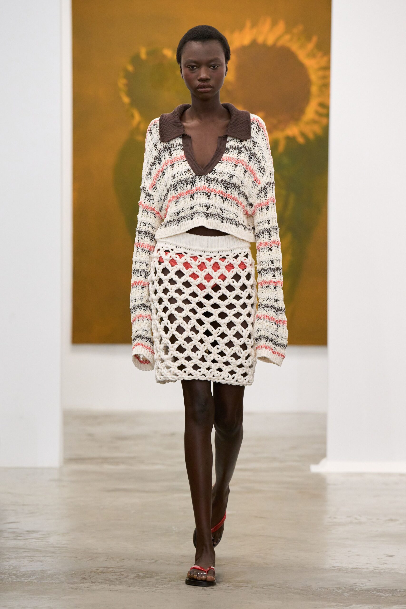 Loose open-knit sweater striped in cream, charcoal and coral, with a deep brown polo collar. Chunky cream macrame mini, diamond netting layered over a coral underpanel. Flat thong sandals with narrow red straps. Walking through a sculpture garden just after the sprinklers stopped.