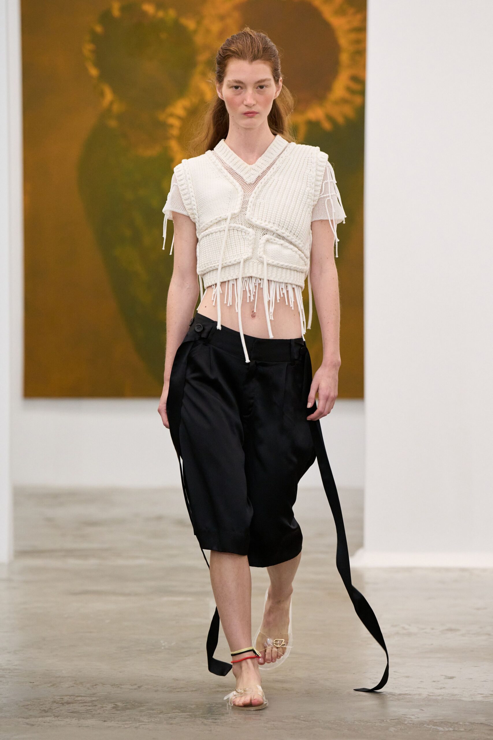 Fine white mesh tee with dangling sleeve ties. Chunky ivory rib knit, paneled and fringed across the bare midriff. Black satin knee-length shorts with oversized side buttons and trailing straps. Clear vinyl sandals with a coral ankle cord. Walking through a museum wing after the air-conditioning finally kicks in.