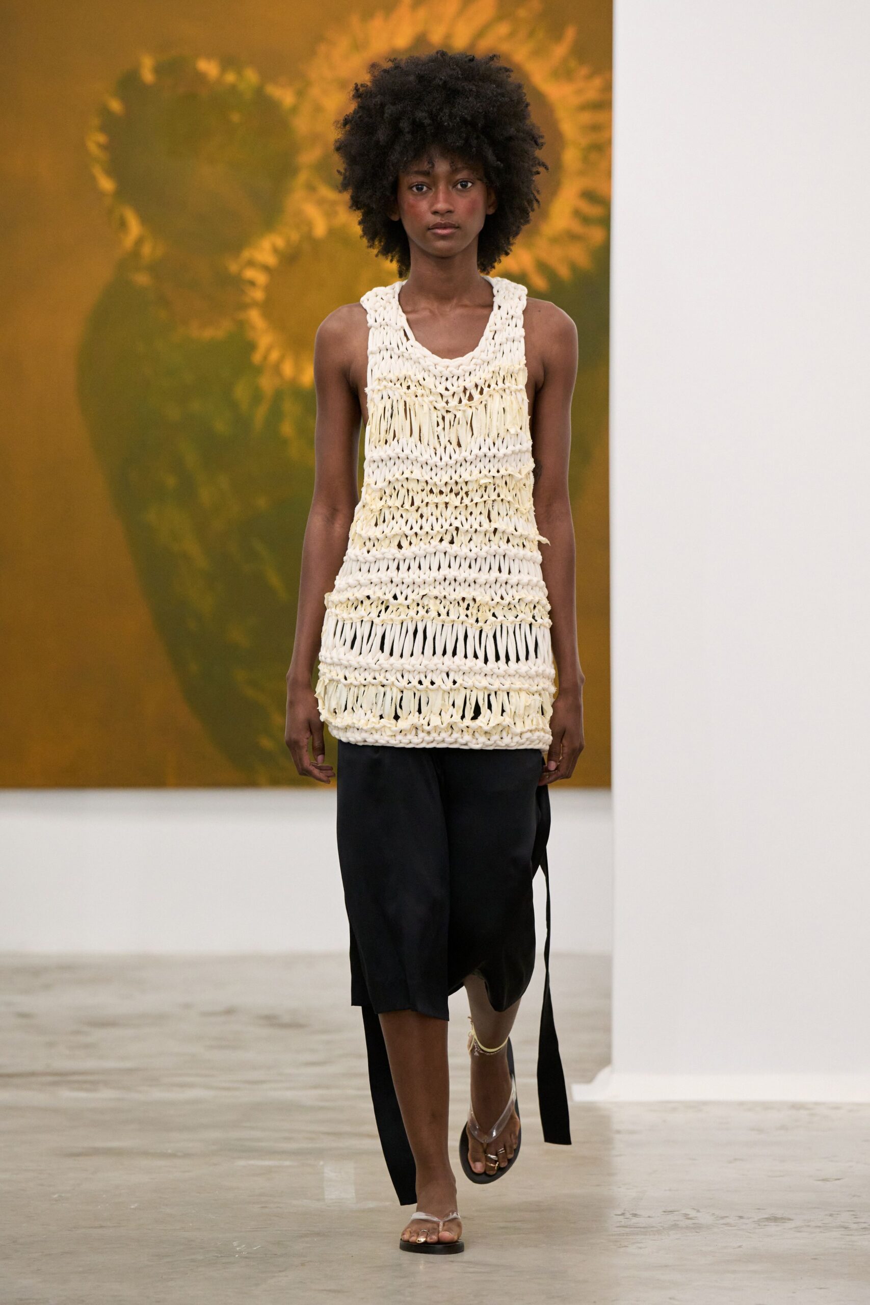 Cream openwork knit tunic in thick looped cord, laddered with long fringed bands. Black wrap skirt hanging below the knee, one ribbon tie trailing loose. Clear thong sandals on flat black soles. Walking through a sculpture court after the rain has dried.