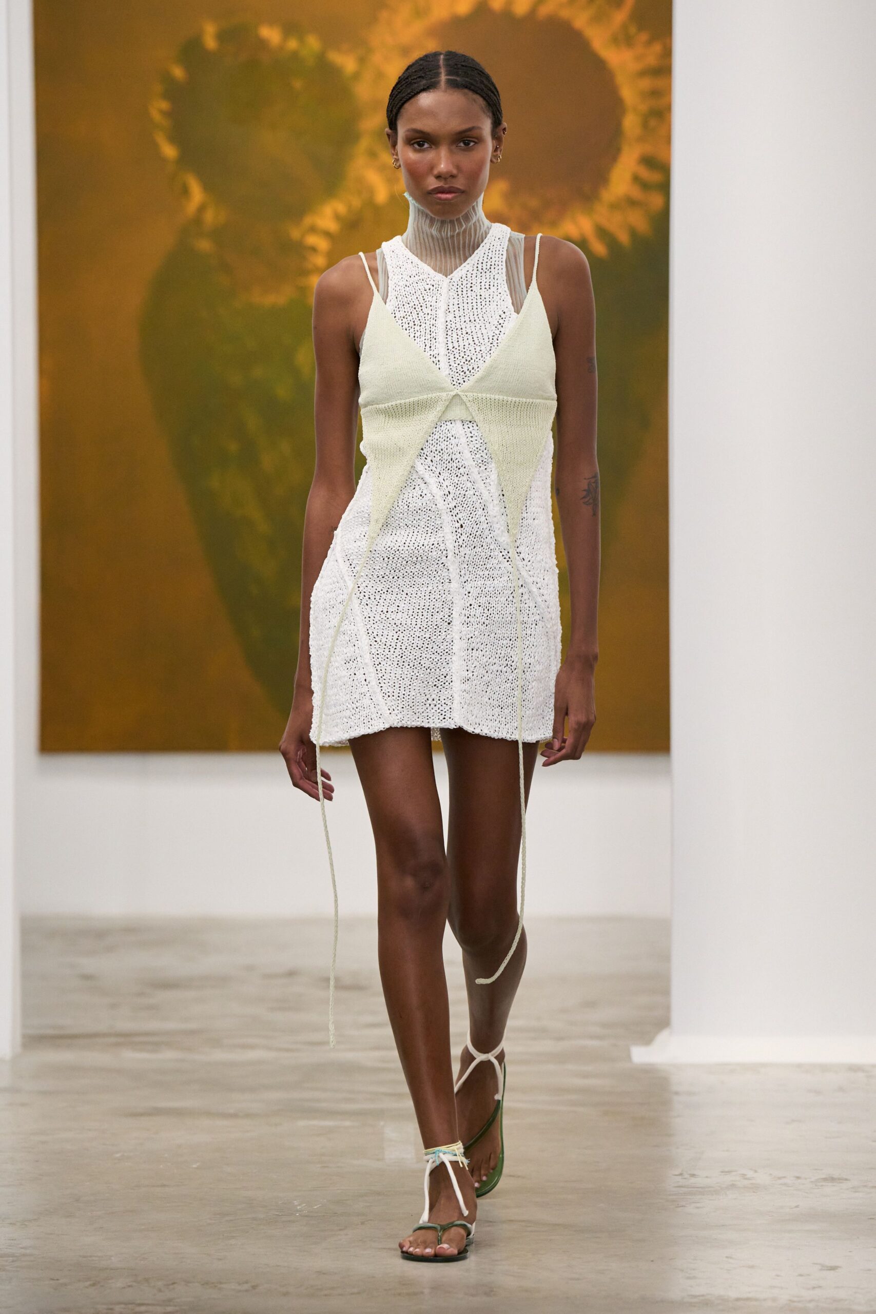 Sheer taupe mesh top with a finely ribbed funnel neck. Open-knit ivory mini, loosely gauged and slightly curling at the hem. Pale citron knit panels crossed into points, cords trailing past the knees. Olive flip-flop sandals laced up with white cord ties. Walking through a museum atrium while the fountains are being switched on.