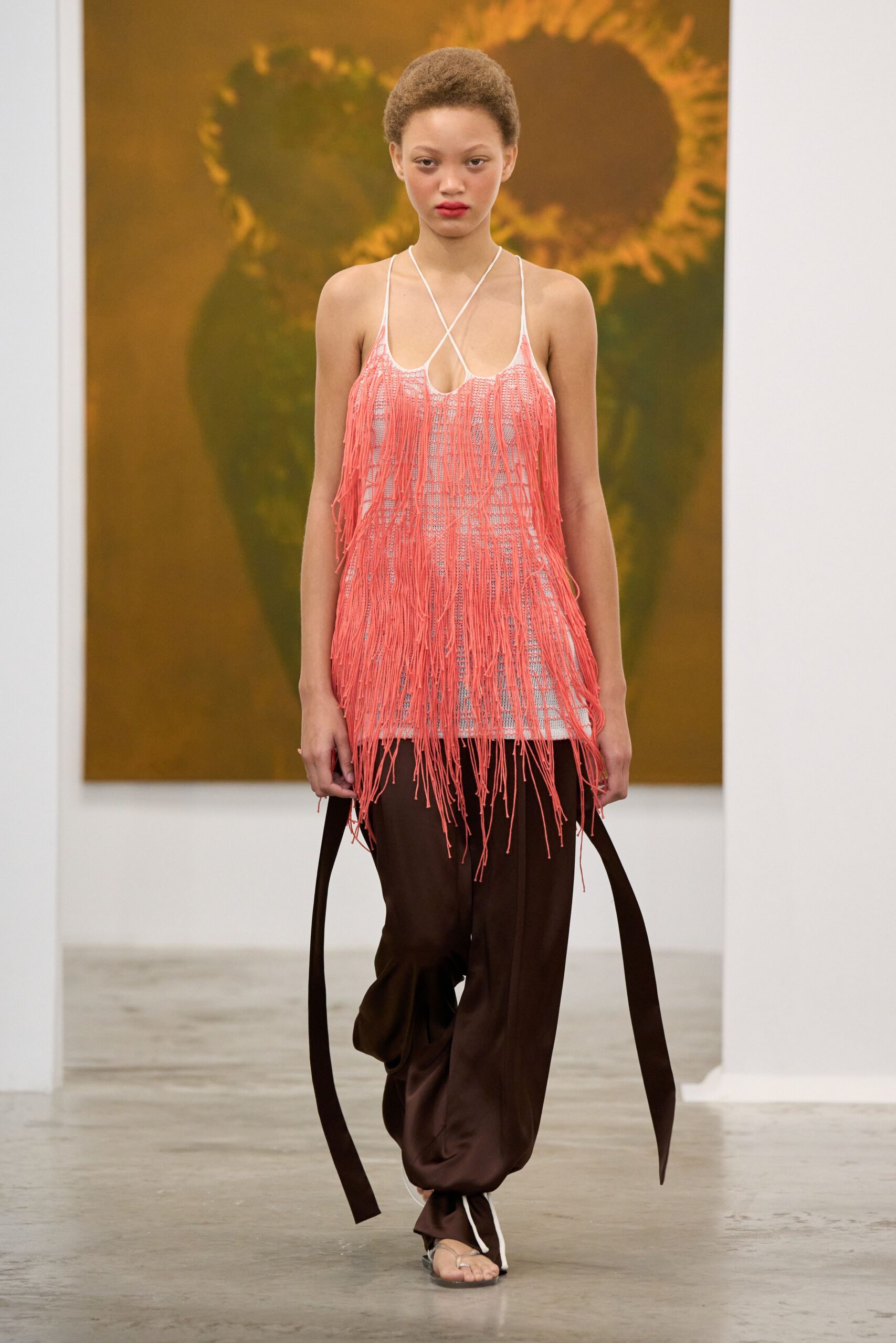 Coral cord fringe over a white open-knit slip, halter strings crossing the chest. Chocolate fluid trousers with extra-long side ties swinging past the calf. Flat thong sandals with a white cord loop at the ankle. Walking barefoot through a beach hotel lobby before breakfast is even cleared.