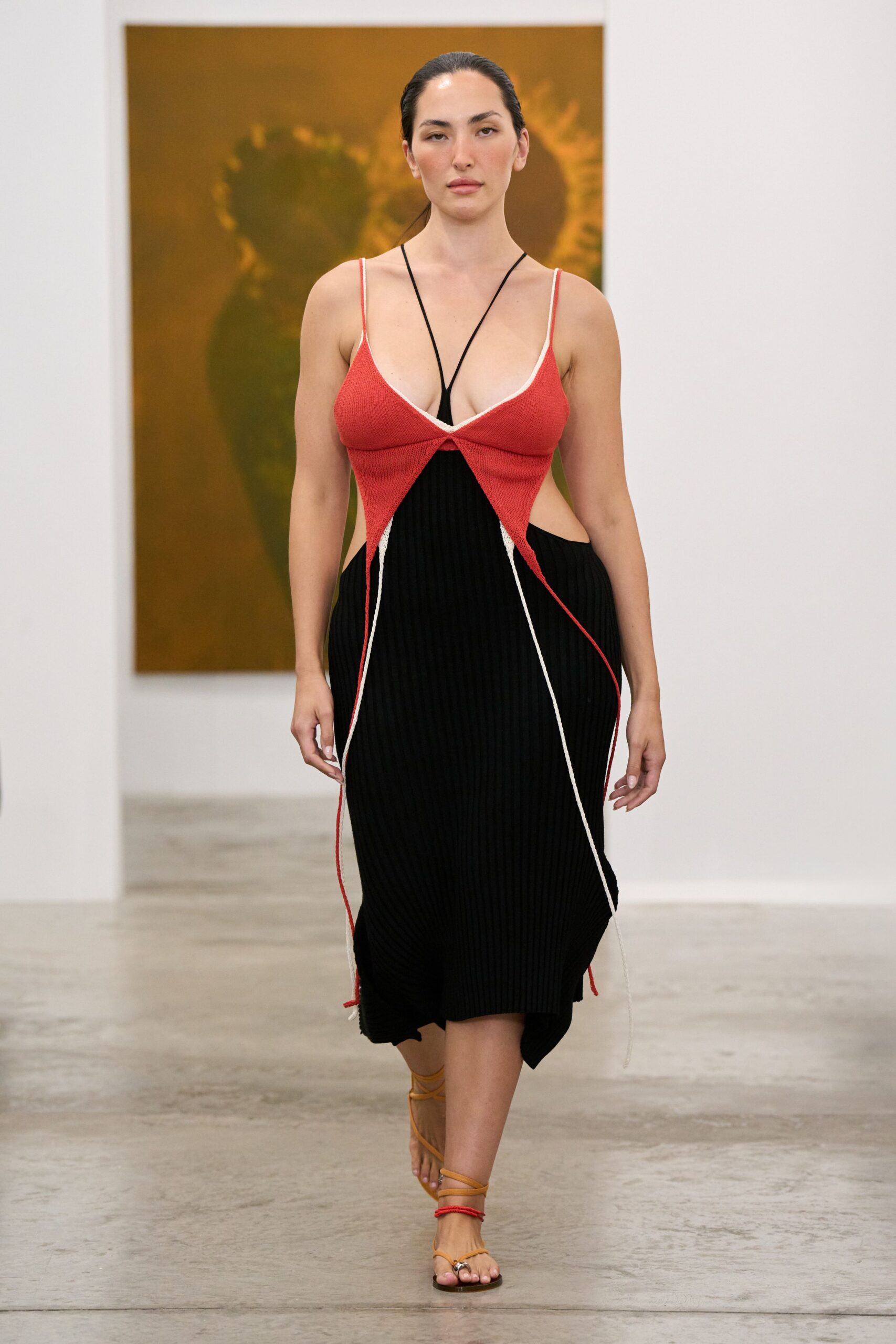Black rib-knit dress with hip cutouts and a handkerchief hem. Coral knit bust overlay, white-edged, ending in long trailing cords. Tan lace-up sandals with one red cord circling the ankle. Walking through a museum atrium after the air-conditioning finally kicks in.