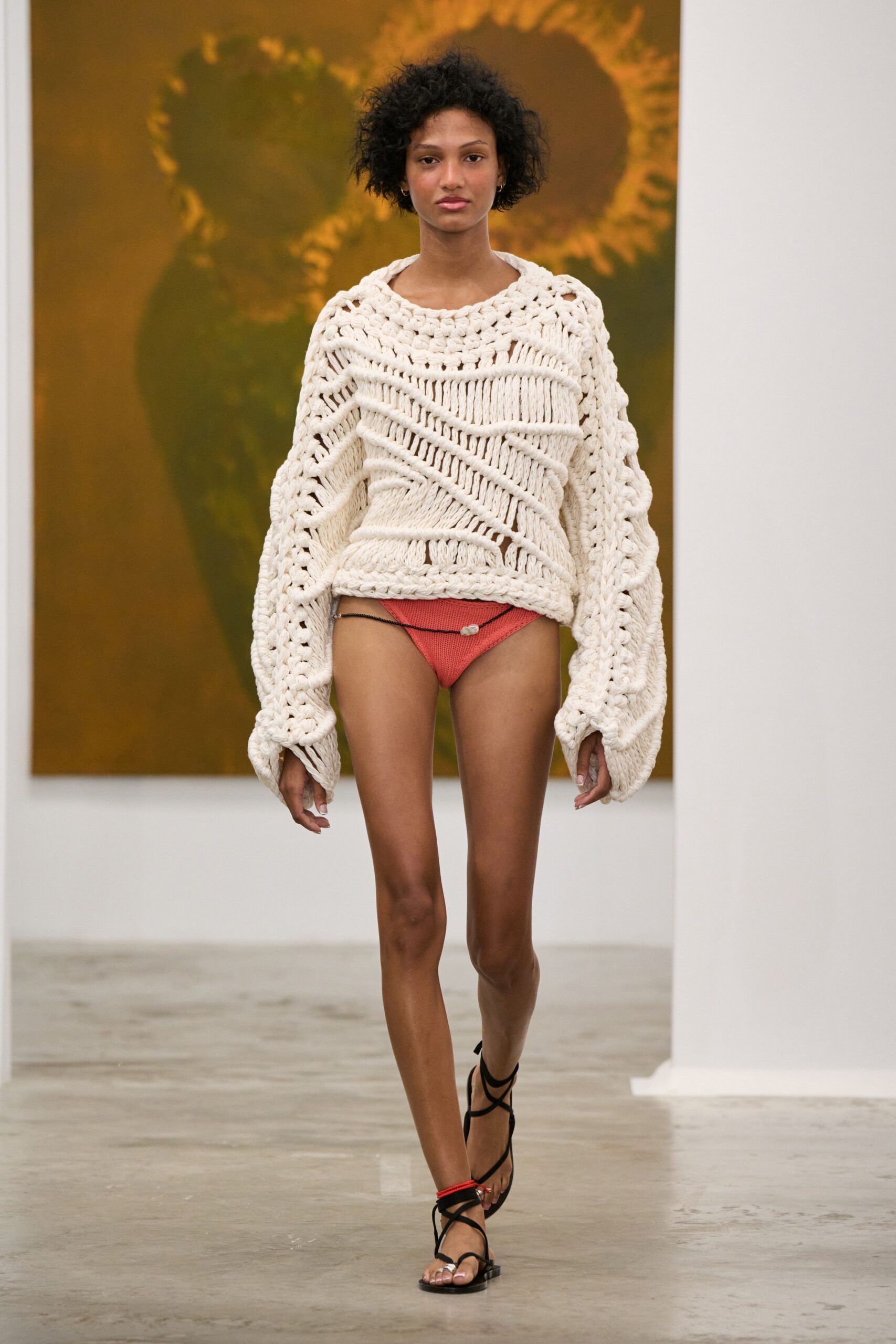 Oversized cream open-knit sweater in thick ropey yarn, sleeves pooling past the hands. Coral ribbed knit bikini bottoms slung from a black cord with one shell bead. Black wrap sandals with a red cord knotted at one ankle. Walking barefoot home from a beach house, sweater grabbed off someone else's chair.