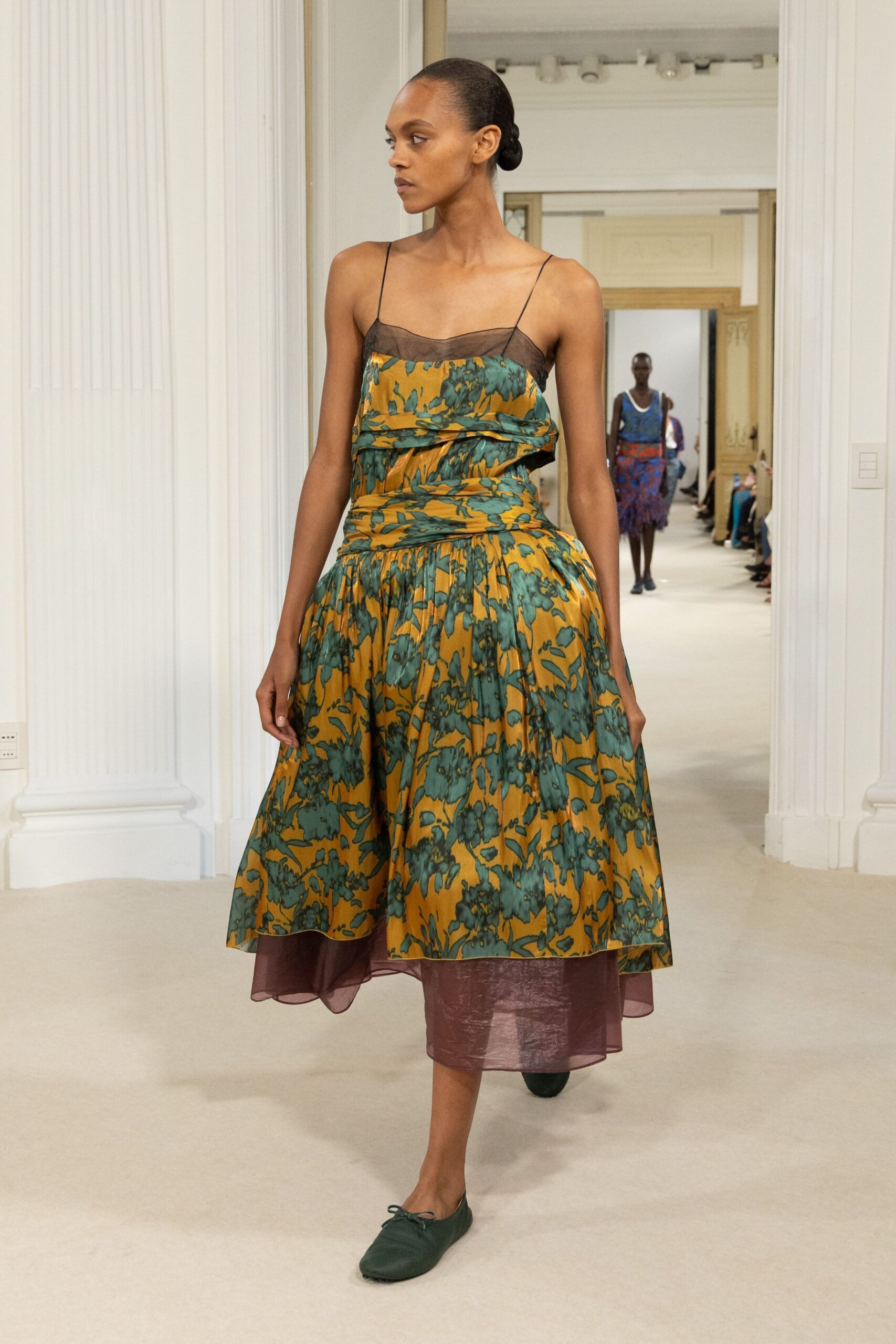 Ochre floral dress with a smoky organza bib and folded waist drape. Dark green ballet flats, each tied with a tiny bow. Crossing a gravel courtyard after the garden wedding has thinned out.