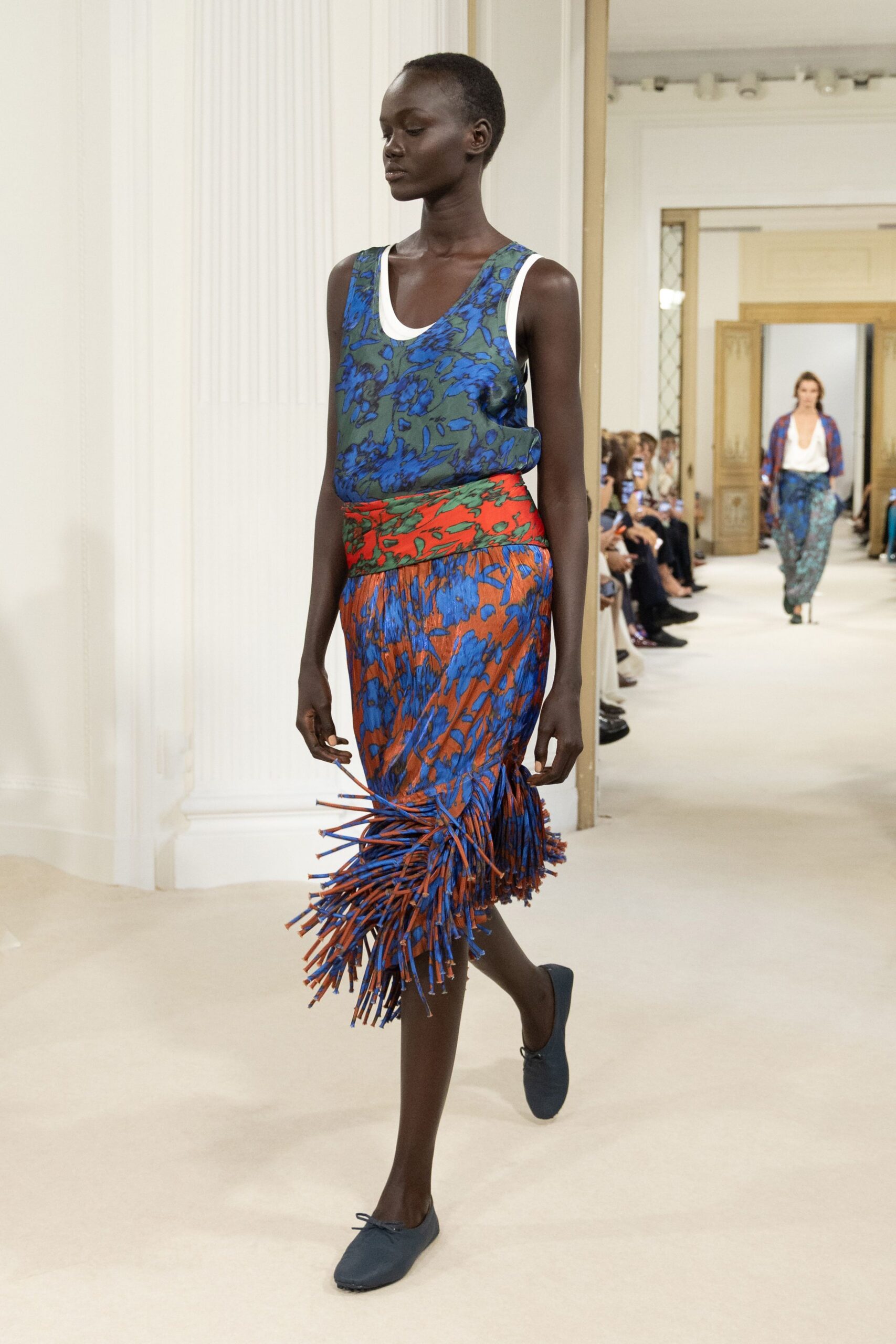 Ink-blue and olive floral tank, white rib binding framing the scoop neck. Rust and cobalt floral wrap skirt, hem exploding into stiff cord fringe. Slate ballet flats with a tiny bow knot at each toe. Crossing a tiled hotel corridor before breakfast, still carrying last night's flowers.