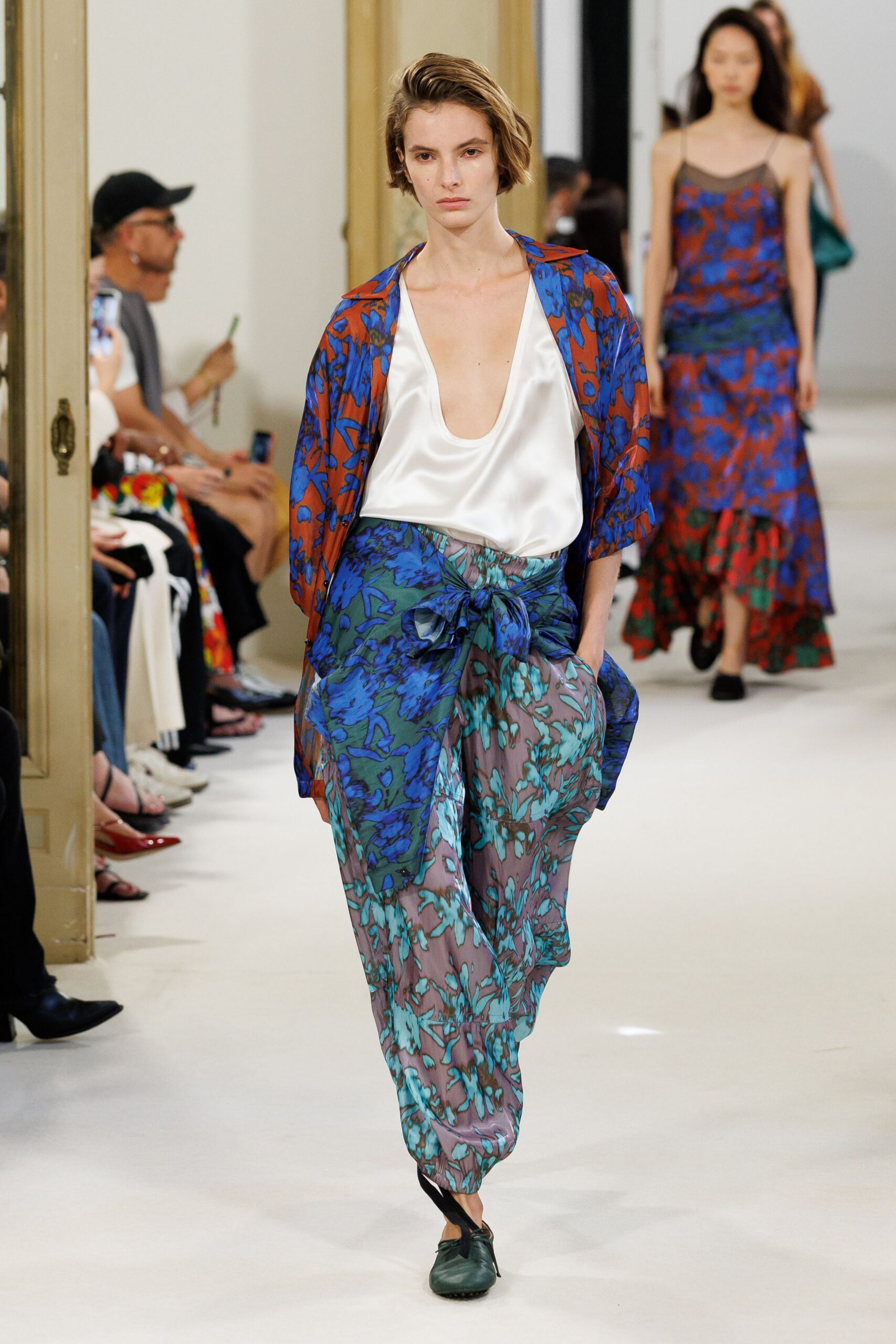 Ivory satin blouse cut with a plunging scooped neckline. Boxy open jacket in rust and cobalt floral print. Loose mauve floral trousers, tied at the waist with a sash knot. Dark green leather flats with black ribbon ties. Walking back from breakfast through a hotel arcade still cool from mopping.