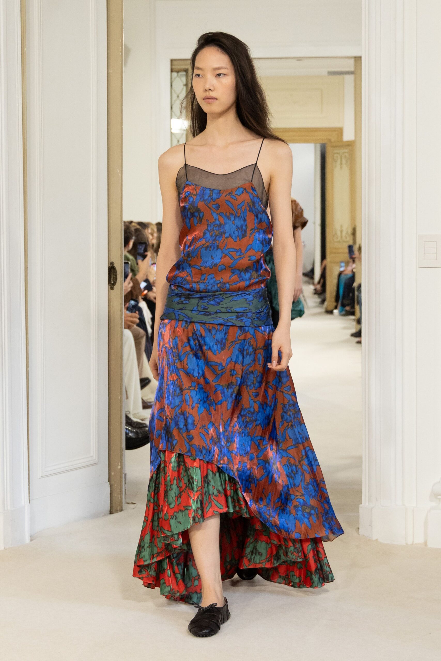 Rust satin slip dress with cobalt florals and a sheer black mesh yoke. Red-and-green floral layer spilling below the uneven handkerchief hem. Black leather ballet flats, drawstring bow slack at the toe. Walking through a hotel corridor after the wedding dinner has thinned out.
