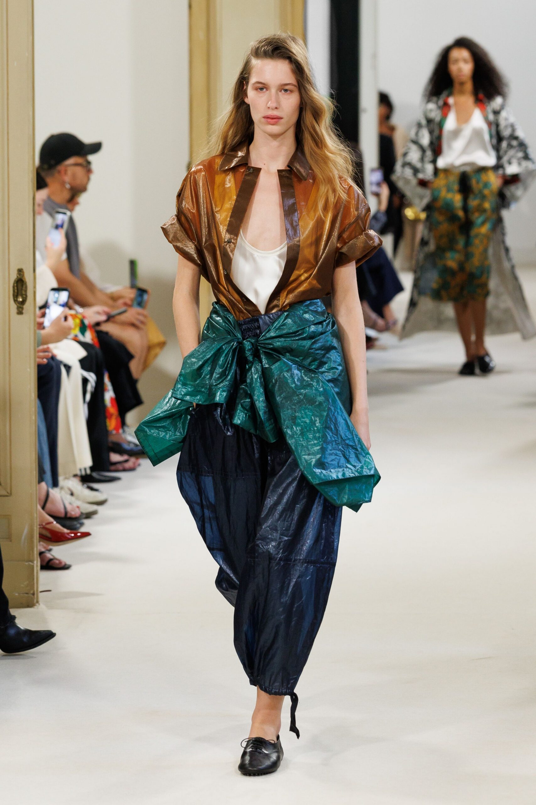 Bronze high-shine blouse, rolled sleeves and dark lacquered placket. White satin camisole cut in a deep scoop. Inky crinkled nylon trousers with a huge teal bow knotted at the waist. Black lace-up flats, one trouser cord dragging near the ankle. For carrying groceries upstairs after rain, still in the clothes from dinner.