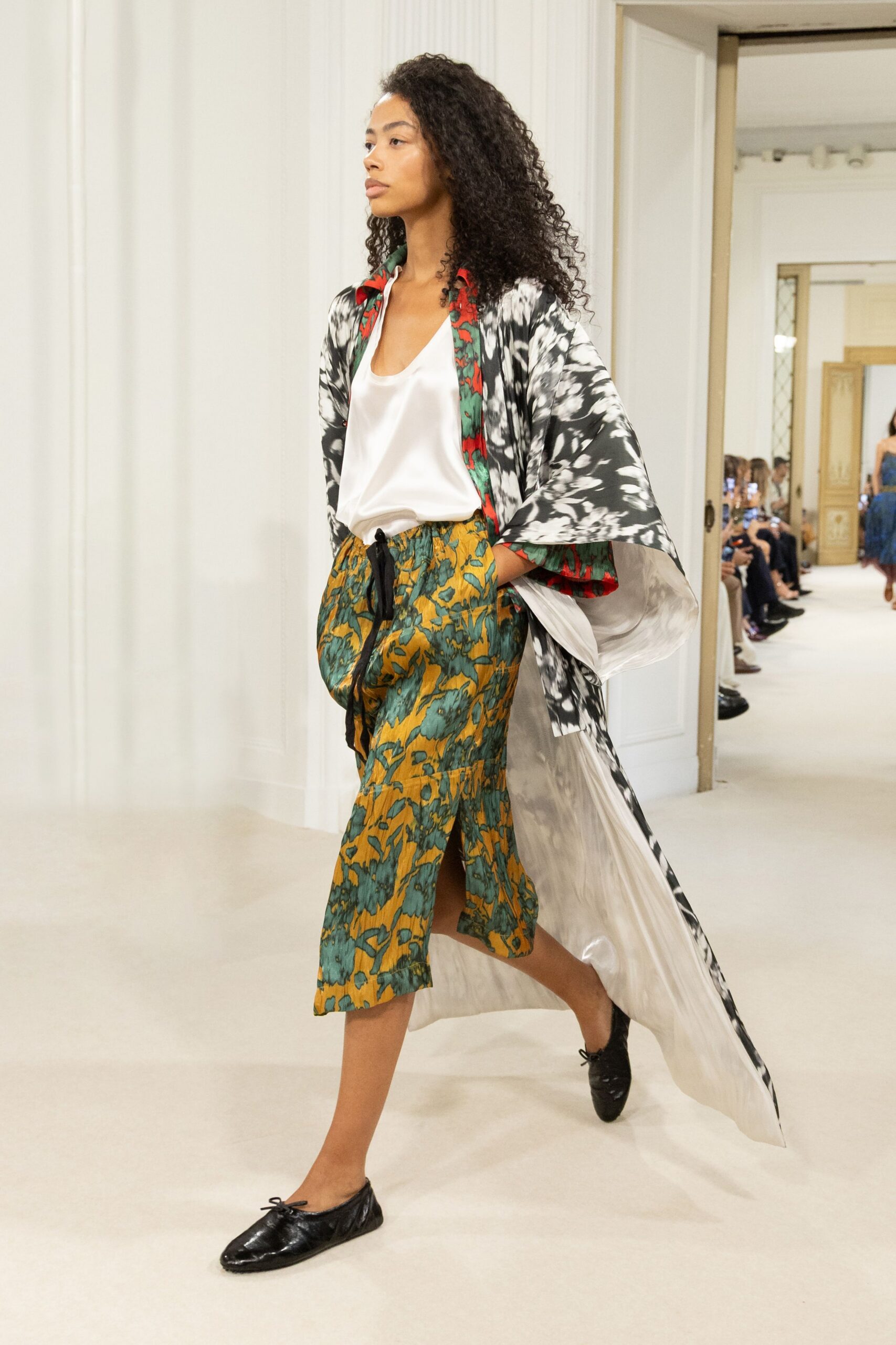 Black-and-white tie-dye robe coat, white satin lining flashing at the stride. White satin tank with a soft scooped neckline. Mustard and teal floral wrap skirt, black tie dangling long at the waist. Black leather ballet flats with tiny bow knots. Walking through a hotel corridor before the wedding guests have come downstairs.