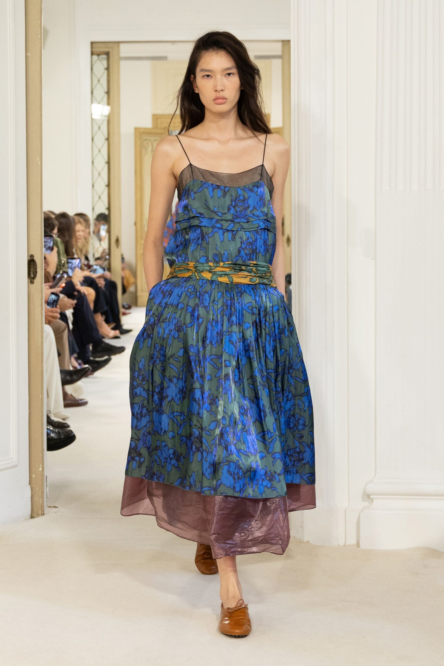 Blue floral dress with a smoky sheer yoke and plum organza underlayer. Twisted ochre-and-green sash cinching the dropped waist in soft folds. Tan leather flats with a knotted tie at the vamp. Walking through a hotel conservatory before the flowers are watered.