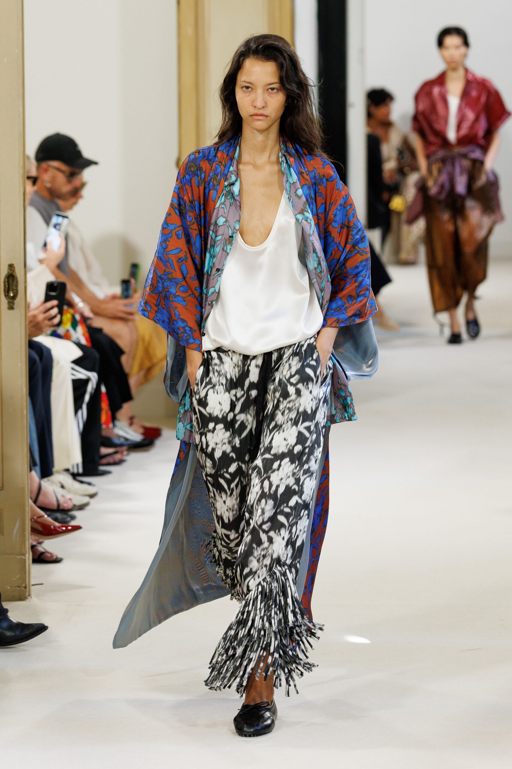 Rust and cobalt floral robe coat, slate lining flashing at the open sides. White satin camisole with a deep scooped neckline. Black-and-white floral trousers, hem breaking into narrow swingy fringe. Black leather flats with rounded toes. Crossing a hotel corridor in the hour after the wedding brunch.