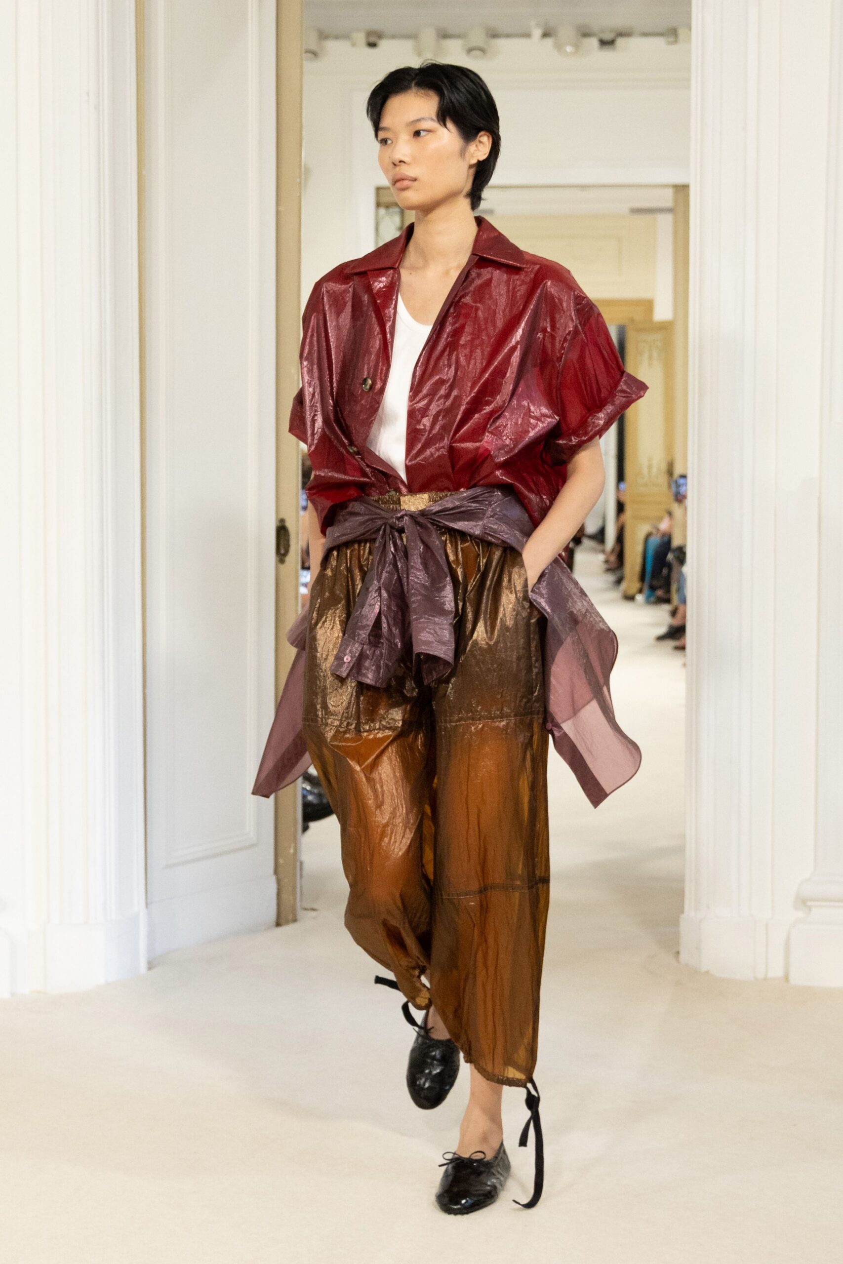 Cranberry crinkled short-sleeve jacket, one dark button sitting off-center. White scoop-neck tank underneath. Plum sheer shirt knotted hard around the waist. Bronze translucent trousers, volume gathered into ankle ties. Black patent lace-up flats. For crossing a hotel lobby with damp hair after the pool.