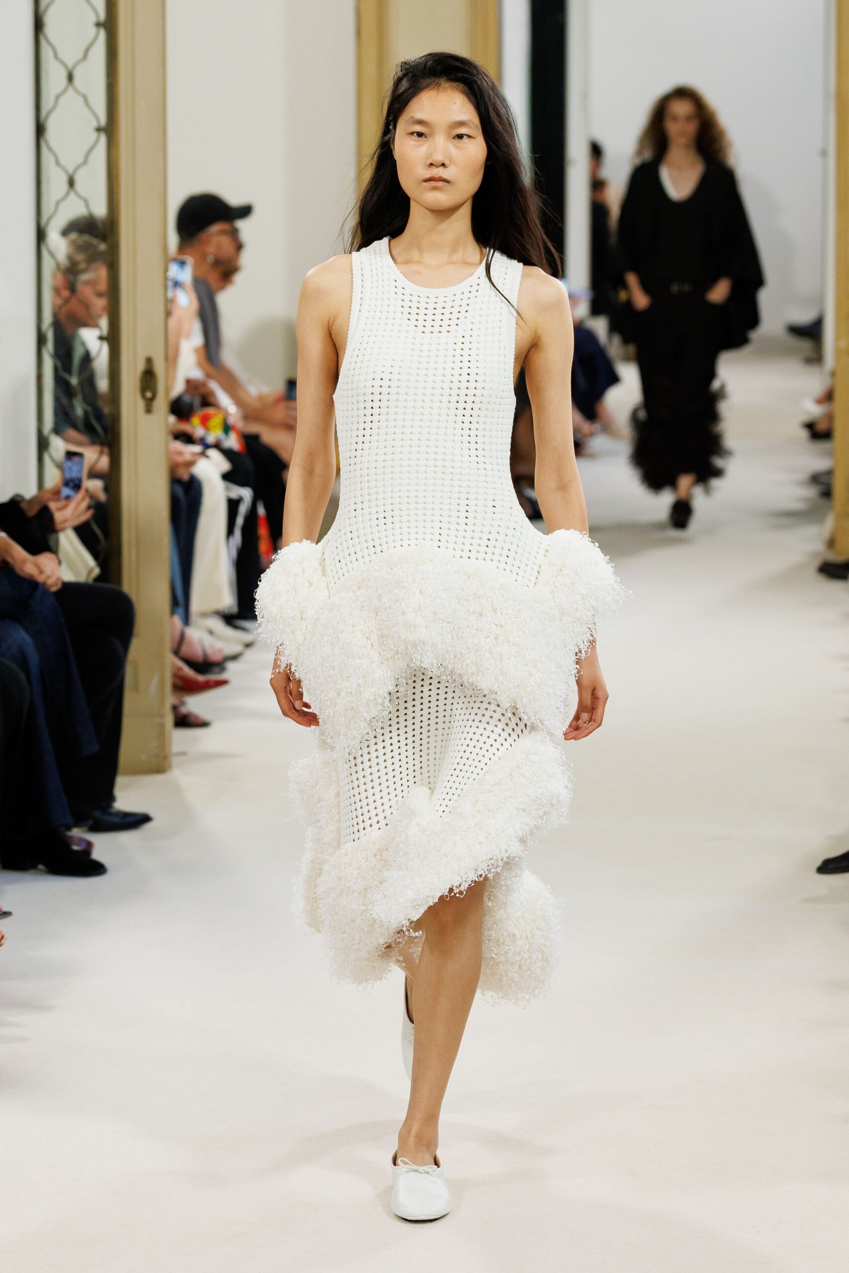 Ivory open-knit tank dress with shaggy bouclé ruffles swelling at hip and hem. White leather flats with a tiny bow pinching the square toe. For crossing a gravel courtyard after the ceremony, bouquet already gone.