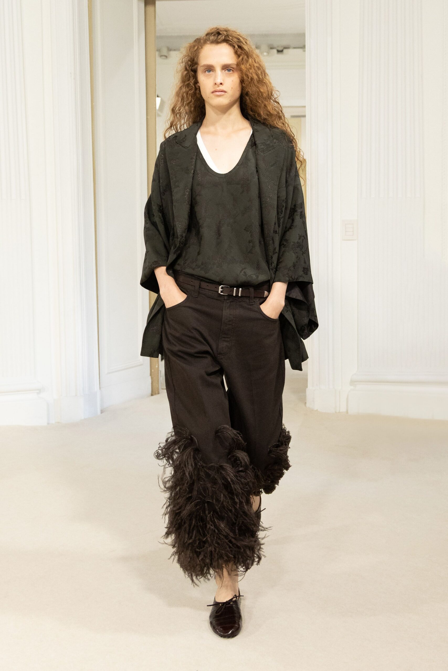 Black floral jacquard jacket cut loose with kimono-like sleeves. Black scoop-neck tank, white underlayer peeking at the collar. Black trousers belted low, shaggy feathered cuffs swallowing the calves. Dark velvet lace-up flats. For crossing a parquet hall while the string quartet is still tuning.