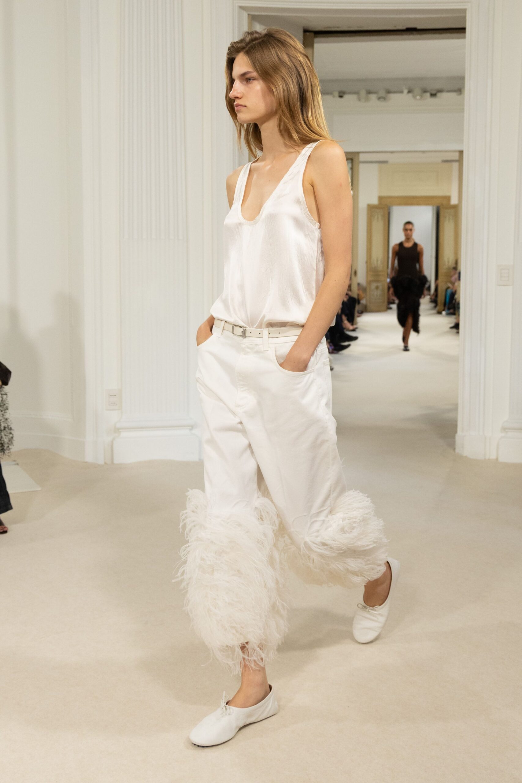 Ivory satin tank, low scoop neck with a liquid drape. Narrow white belt with a small square buckle. Cream cropped trousers, shaggy feather bands swelling around both calves. Soft white leather flats, tied with tiny front bows. Crossing a hushed hotel corridor before someone opens the ballroom doors.