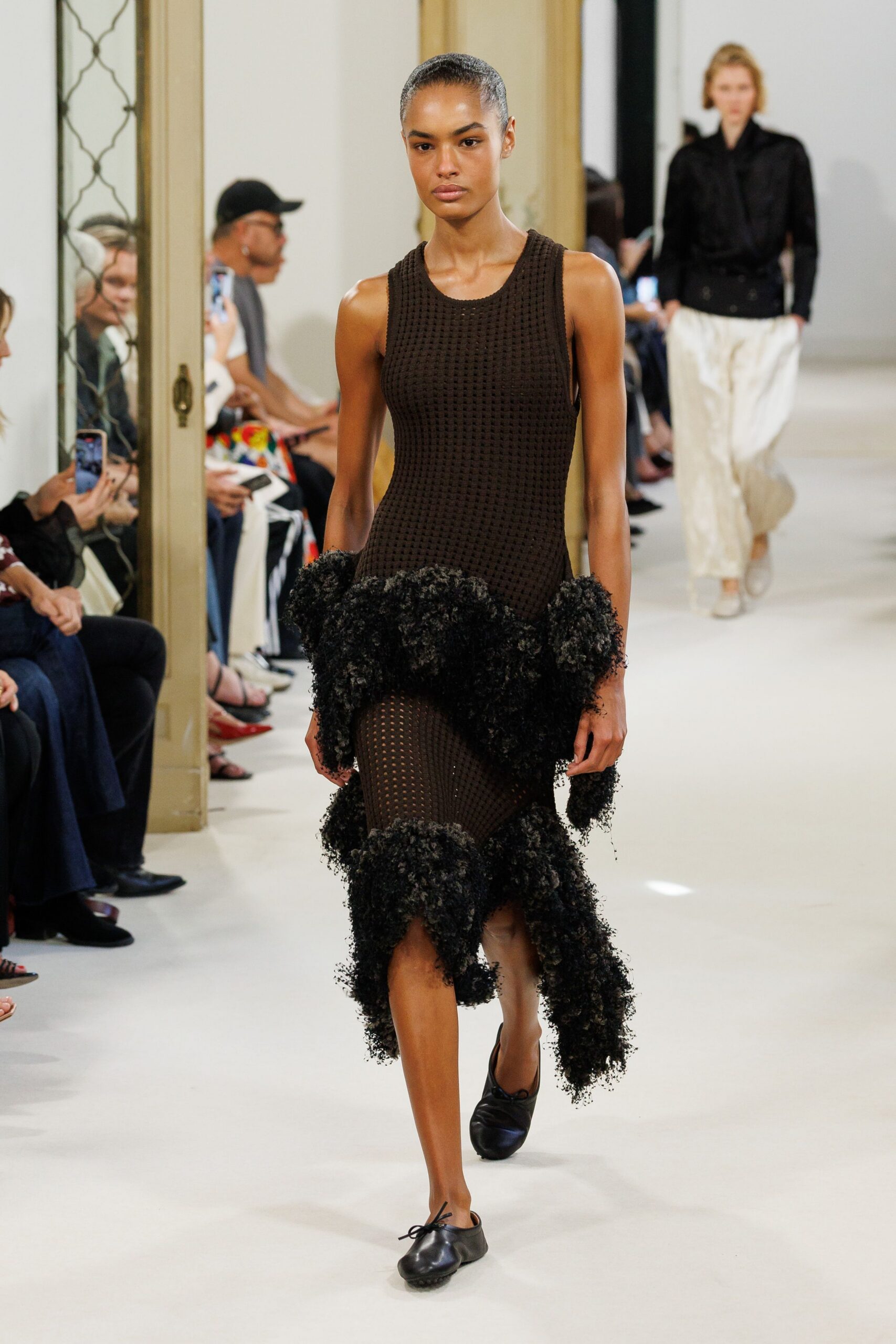 Dark espresso open-grid knit tank dress with shaggy bouclé flounces at hip and hem. Black leather ballet flats with a tiny bow and gathered toe. Crossing a hotel corridor before breakfast, carrying yesterday into the light.