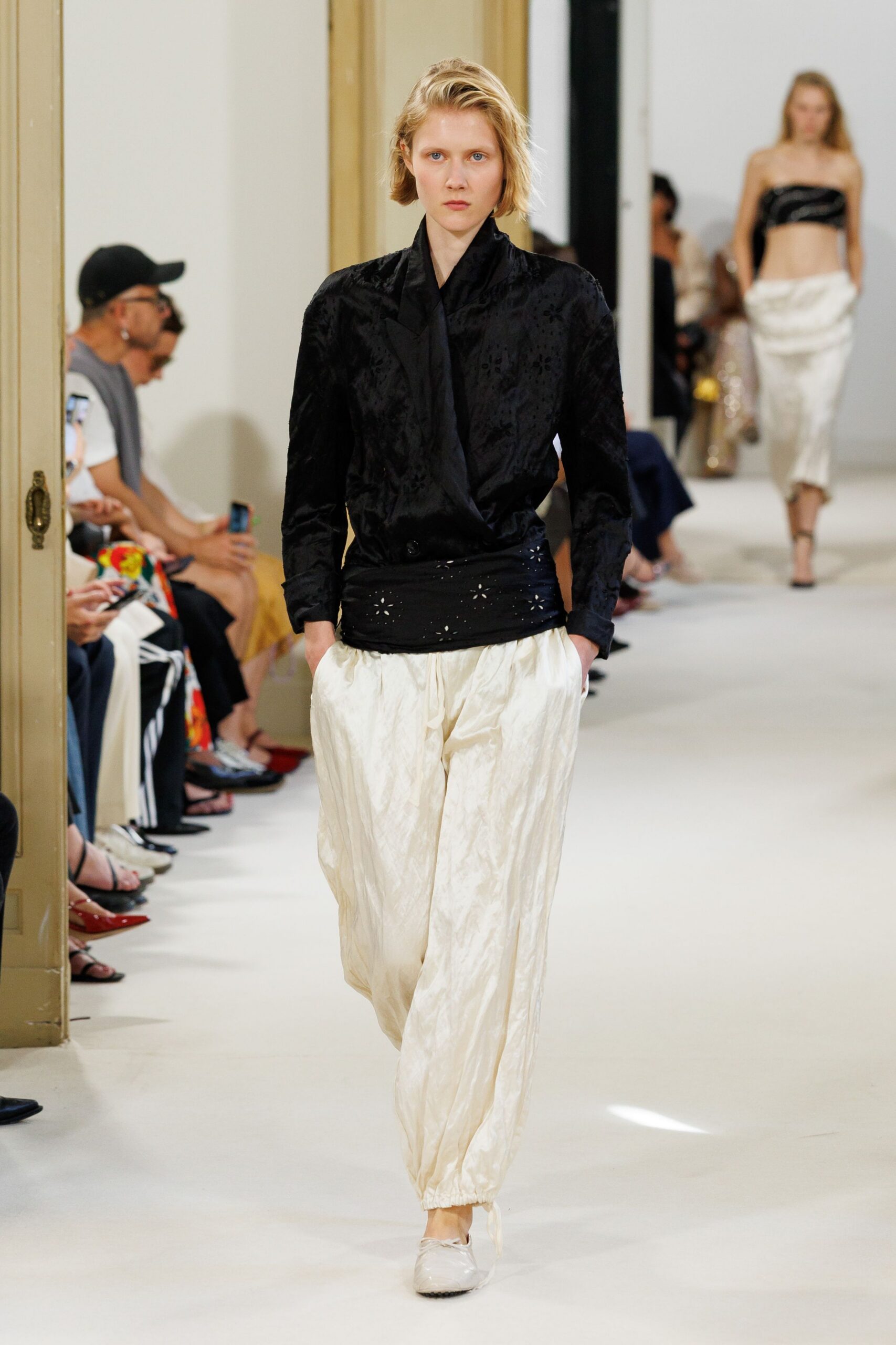 Black floral jacquard jacket with padded shoulders and a shawl-wrap front. Black sash waist wrap pricked with tiny eyelet flowers. Cream crinkled trousers, gathered tight at the ankle. White rounded flats. Crossing a tiled courtyard after midnight rain, still carrying dinner in your posture.
