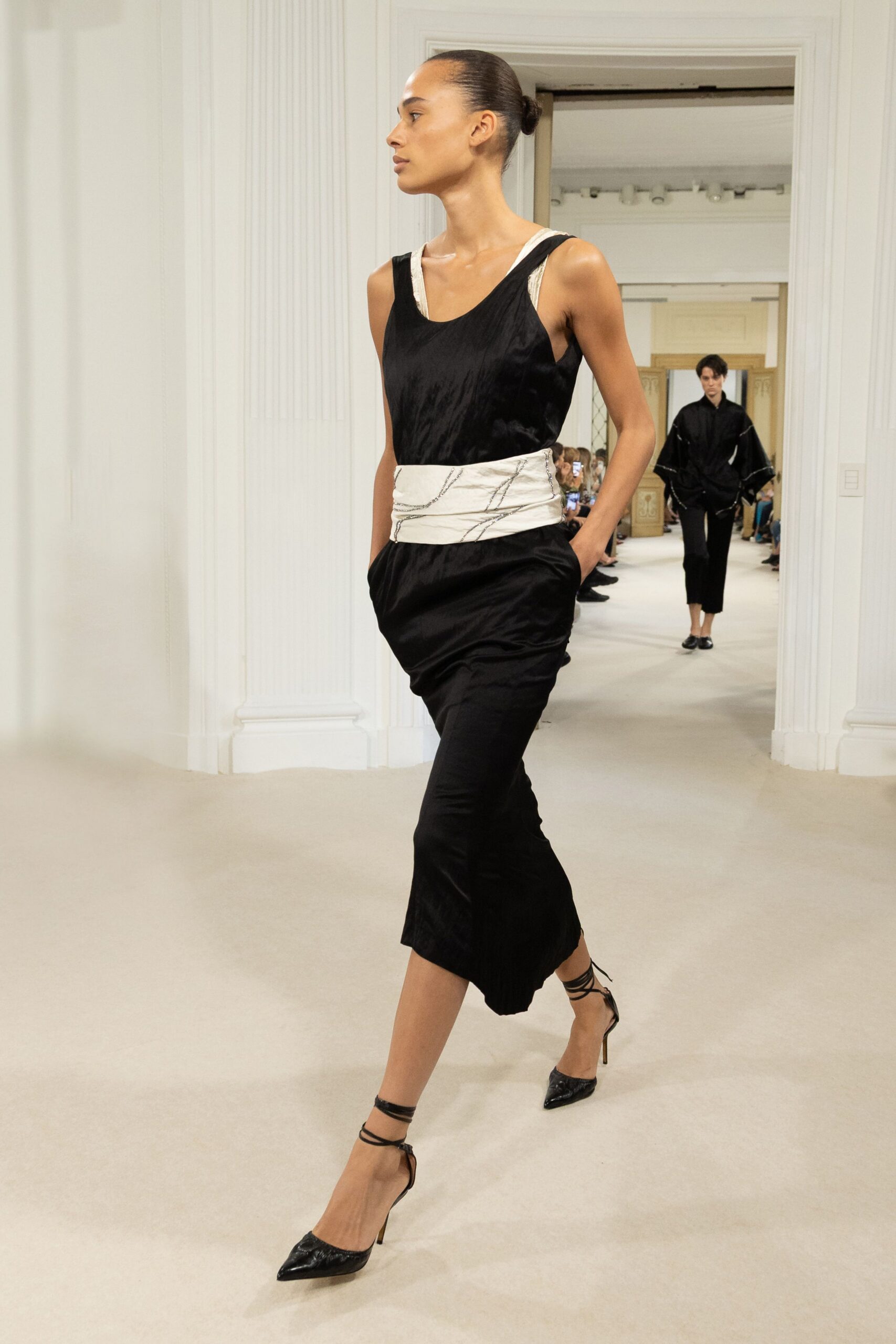 Black satin slip dress, cut low at the armhole over white straps. Wide ivory sash, scribbled with dark beaded lines. Black pointed pumps with thin ankle ties trailing loose. Crossing a marble foyer before the string quartet starts.