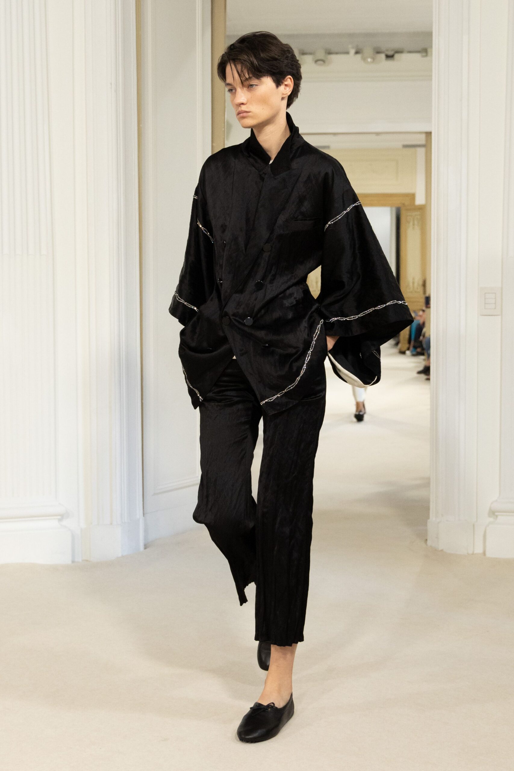 Black satin jacket with kimono sleeves and white cord tracing the seams. Black crinkled trousers, narrowly cut and cropped above the ankle. Black leather ballet flats with a tiny bow at the vamp. Walking the hotel corridor after midnight with your shoes still on.