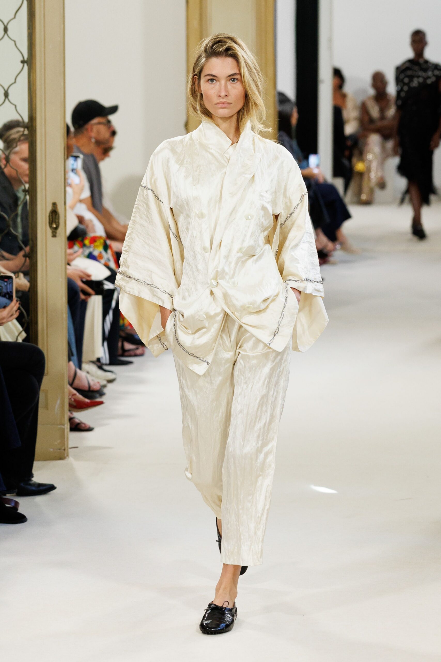 Ivory crinkled satin jacket with kimono sleeves and loose silver whipstitching. Matching cropped trousers in the same crushed sheen. Black patent lace-up flats, softly collapsed at the toe. For opening the balcony doors after a wedding everyone else already left.