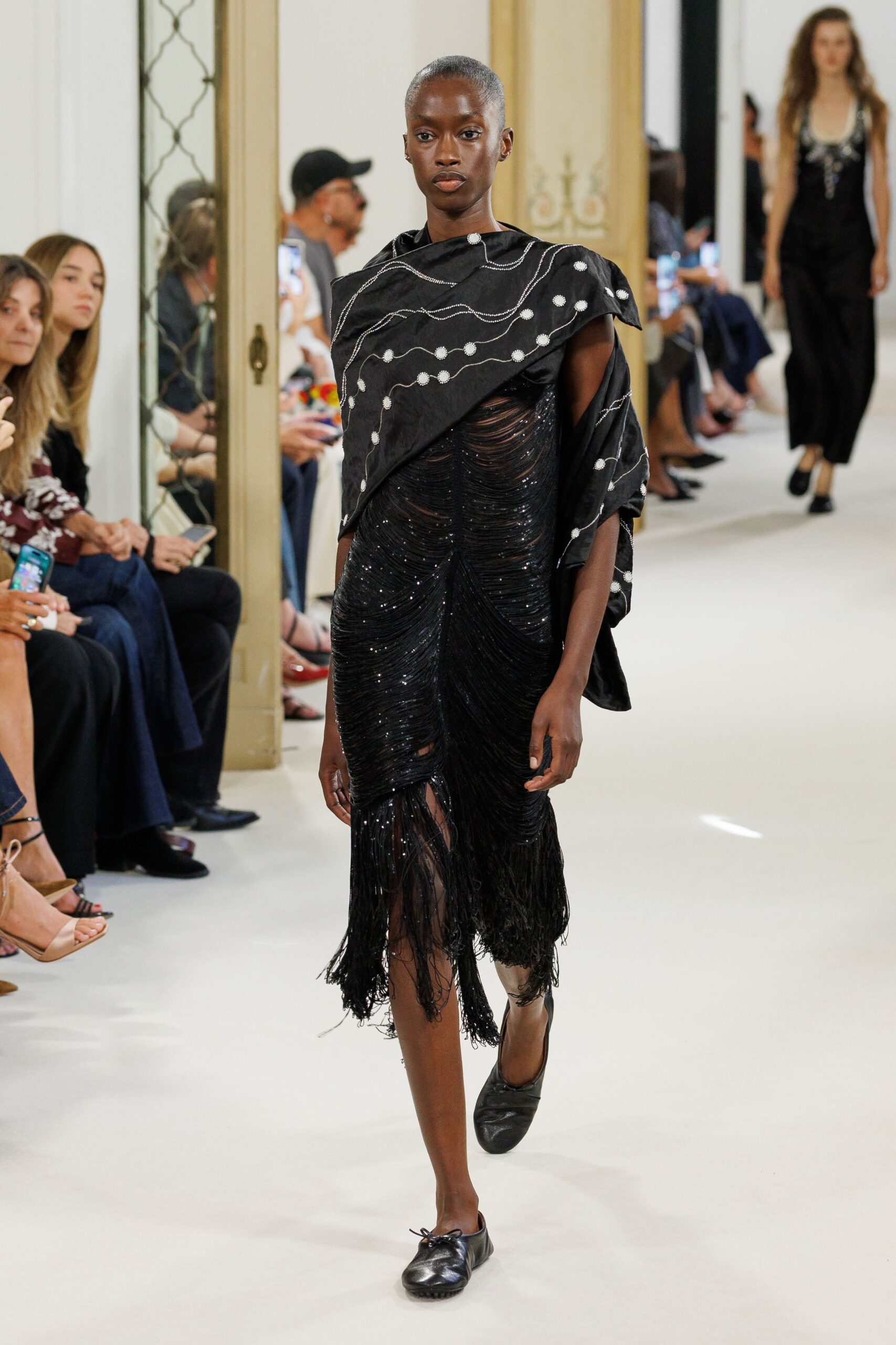 Black wrap embroidered with white dotted squiggles, folded into a skewed shoulder cape. Black beaded fringe dress, densely ruched into scalloped swags and a ragged hem. Soft black leather flats, bow knot loosening at one toe. Crossing a hotel ballroom after midnight while the band packs up.