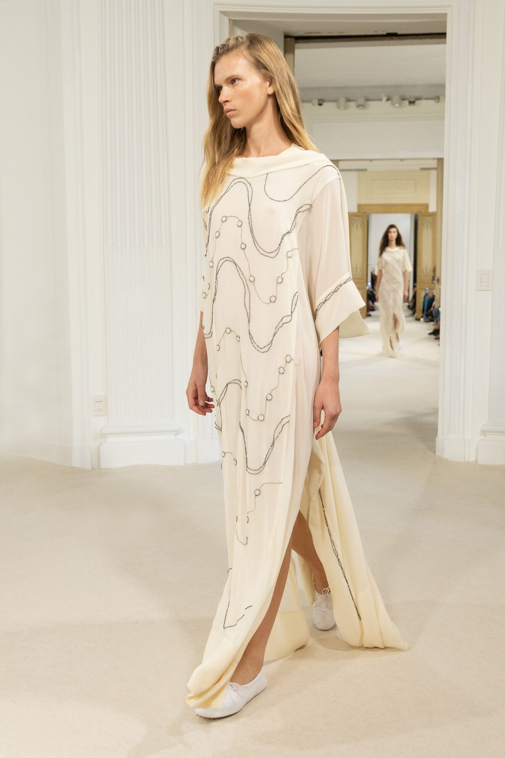 Ivory silk caftan with a folded boatneck and loose kimono sleeves. Black beaded squiggles and tiny ring motifs wandering across the cloth. White lace-up flats, almost hidden by the pooling hem slit. Crossing a silent museum atrium before the doors open to the public.
