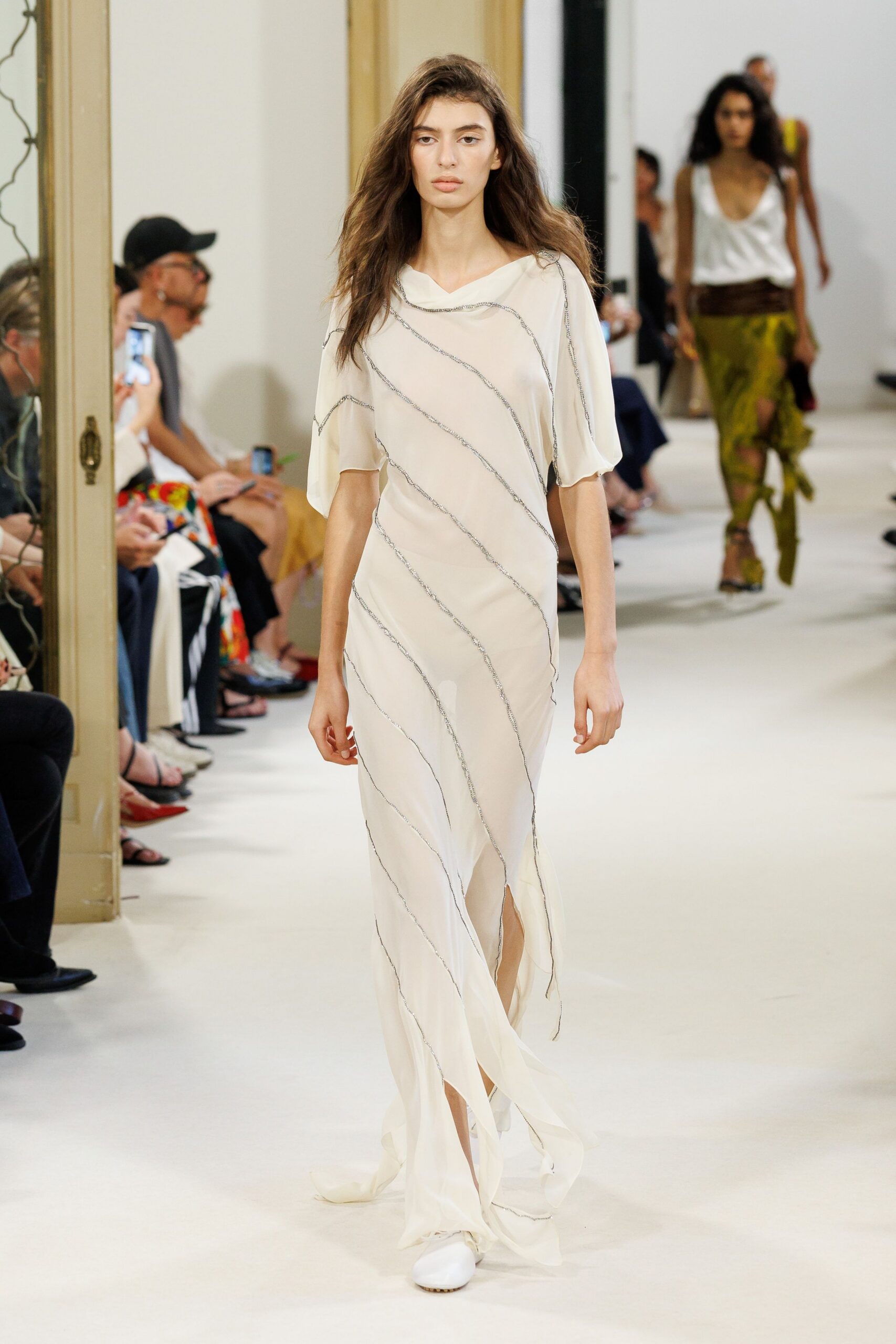 Ivory chiffon column gown with silver-beaded seams wandering diagonally. White rounded flats peeking from the torn front slit. Crossing a hotel corridor barefoot in your head, heels already in the bag.