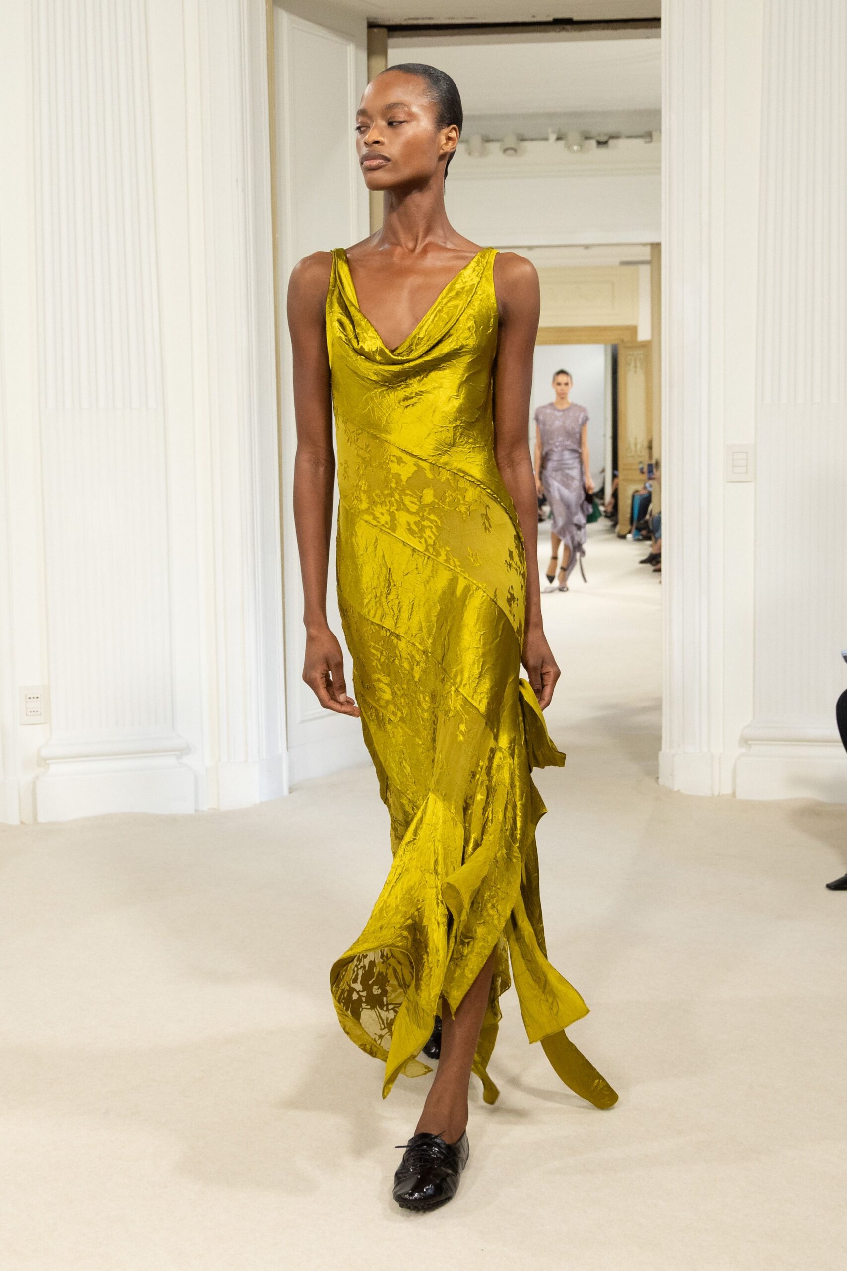 Chartreuse hammered satin gown with a draped cowl and spiraling seamwork. Black crinkled leather flats, laces knotted low over the instep. Crossing a marble foyer while the florist is still sweeping stems.