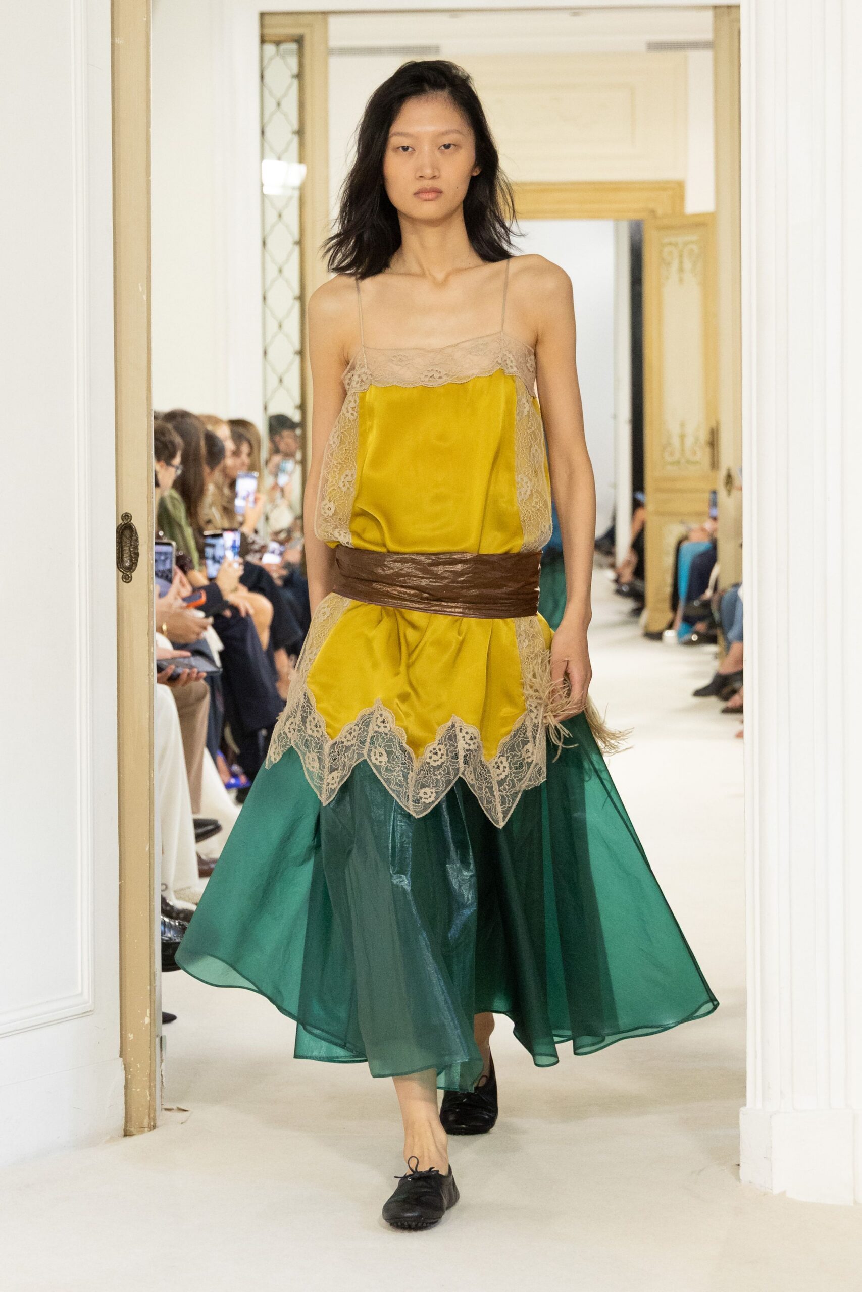 Marigold satin slip dress, beige lace scalloping across the bust and hem. Wide crinkled brown sash cinched low around the hips. Bottle-green organza overskirt, sheer volume floating away from the knees. Black leather ballet flats with a short bow knot. Crossing a hotel corridor after midnight with flowers already shedding.