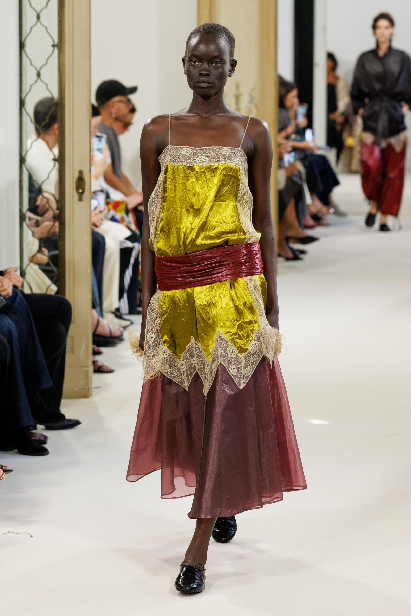 Chartreuse crinkled satin slip dress veiled with cream floral lace. Oxblood glossy sash wound low and tight through the waist. Sheer plum organza overskirt falling from under the jagged lace hem. Black patent flats, softly collapsed across the vamp. For crossing a hotel ballroom after midnight with petals stuck to the floor.
