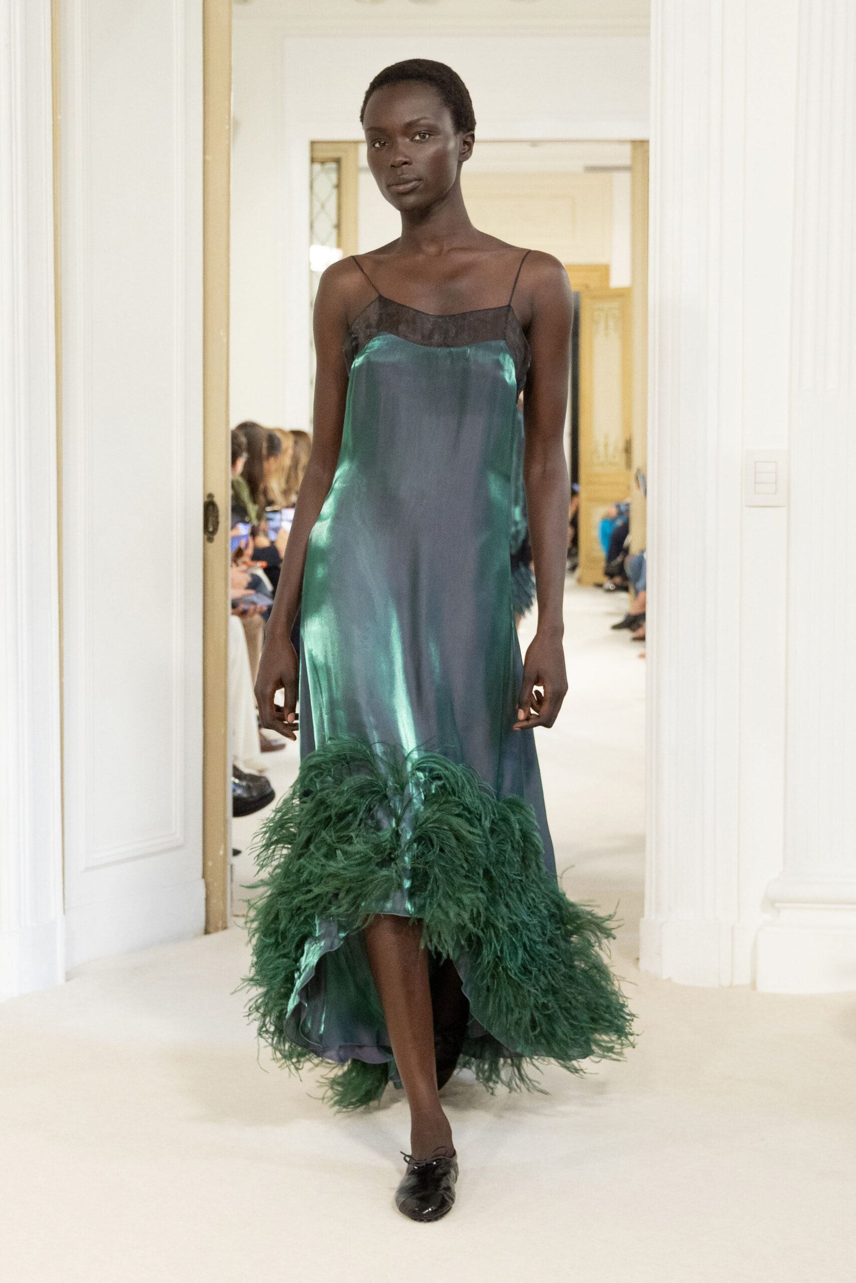 Petrol satin slip dress with a sheer black yoke and trailing ostrich-feather hem. Black patent lace-up flats, square at the toe. Crossing a hotel ballroom after midnight, feathers still stirring the waxed floor.