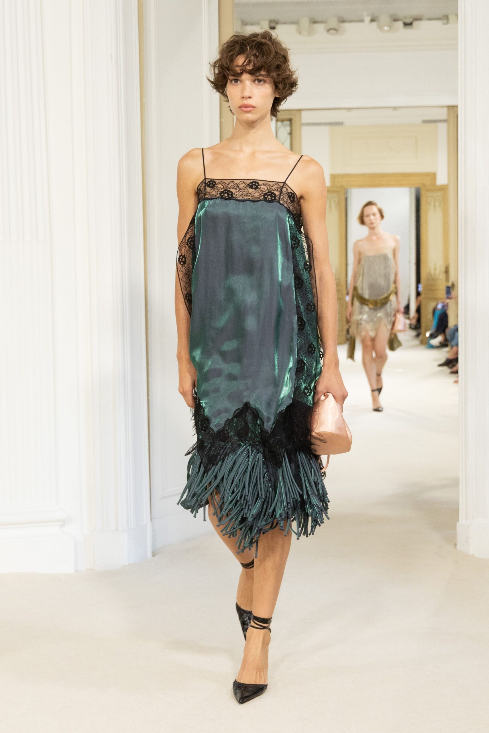Petrol satin slip dress edged in black floral lace, rubbery fringe swinging below. Blush satin pouch tucked soft in one hand. Black pointed pumps with wraparound ankle ties. Crossing a hotel corridor after midnight with flowers already shedding.
