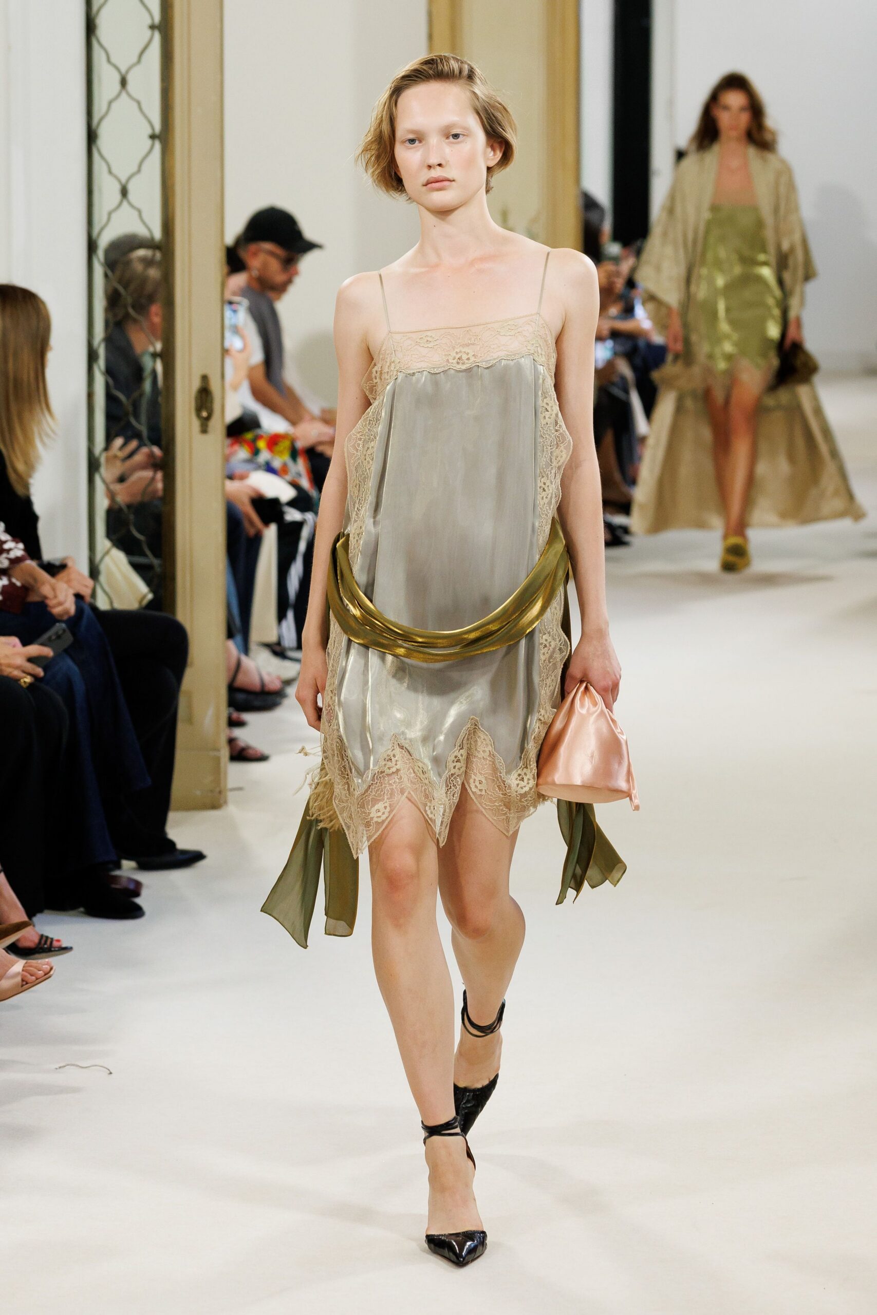 Pewter satin slip dress with ecru lace edging and a jagged handkerchief hem. Olive chiffon scarf slung low, pinned by a molten gold crescent. Blush satin drawstring pouch, softly wrinkled in the hand. Black patent slingbacks with thin straps winding the ankle. Hotel corridor at noon, still carrying last night's perfume.