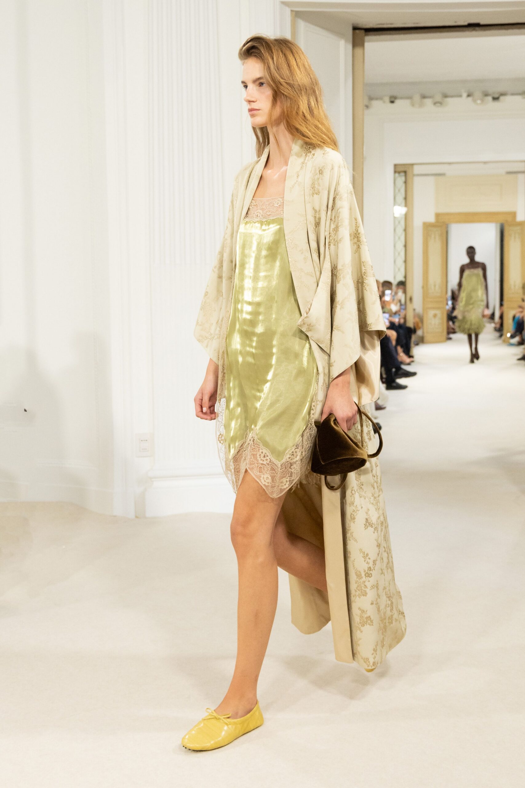 Champagne floral jacquard robe coat with wide kimono sleeves. Lemon satin slip dress, scalloped lace edging the neckline and hem. Crushed brown velvet pouch collapsing around a thin loop handle. Butter yellow flats with a tiny bow at the throat. Morning after a hotel wedding, carrying your shoes back downstairs.