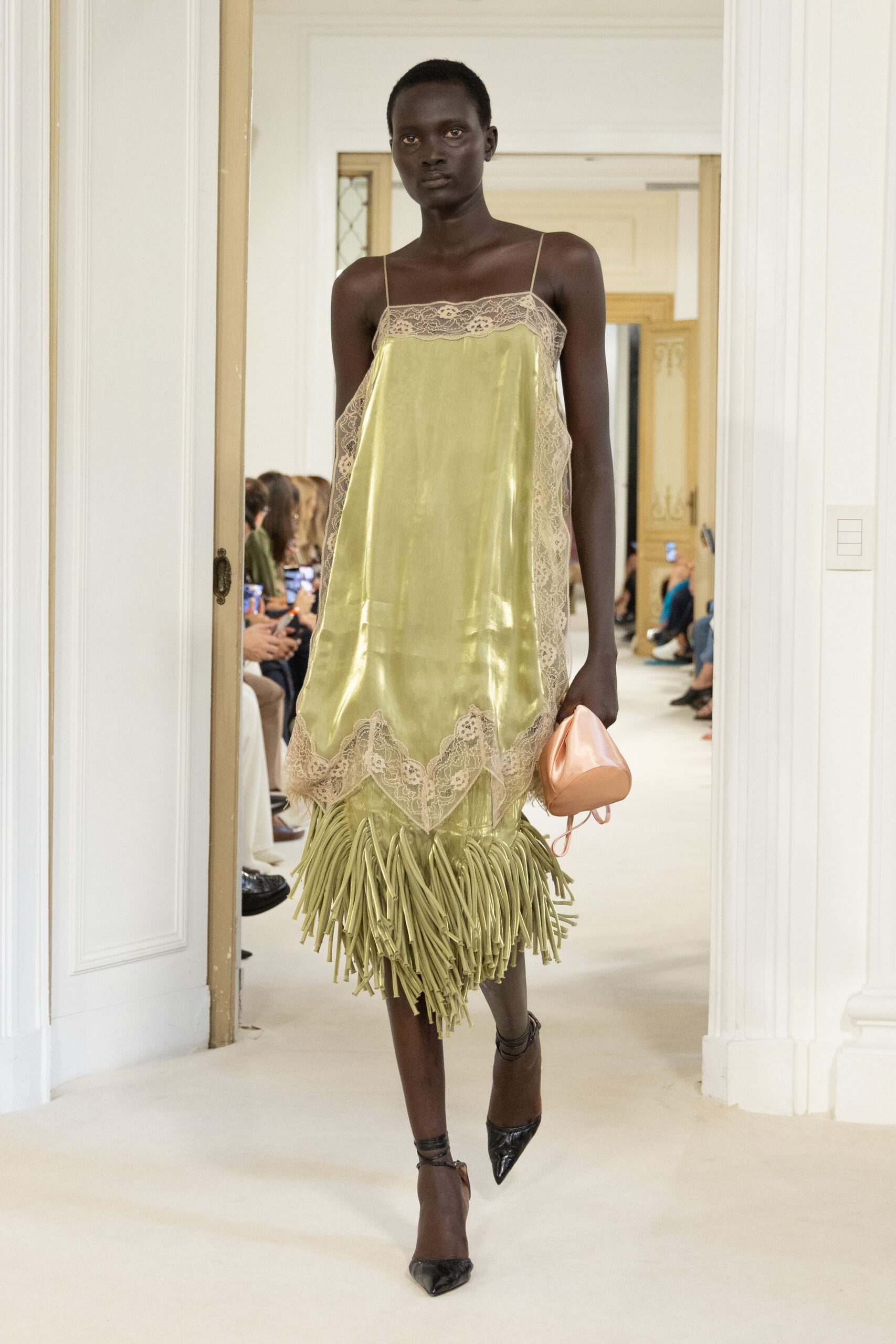 Chartreuse satin slip dress, ecru lace paneling and a long cut-fringe hem. Peach satin drawstring pouch, softly collapsed in the hand. Black pointed pumps with thin wraparound ankle straps. Walking through a grand hotel corridor before anyone has opened the ballroom.