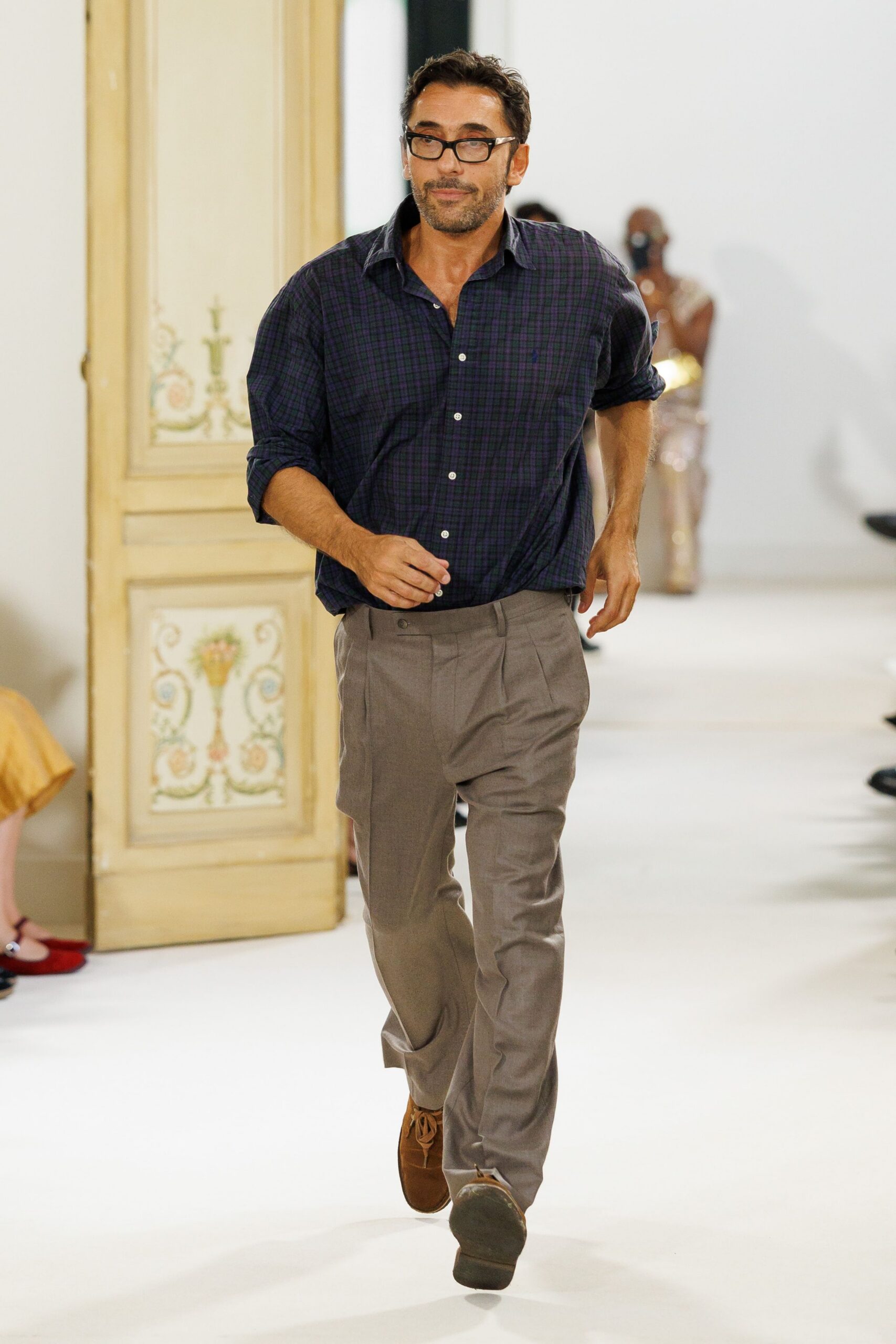 Black rectangular frames with thick temples. Indigo-and-plum tartan button-down, sleeves pushed to the forearm. Taupe flat-front trousers with a sharp crease and slanted pockets. Tobacco suede lace-ups, softly rounded at the toe. Parent-teacher conference, then dinner somewhere with paper tablecloths.
