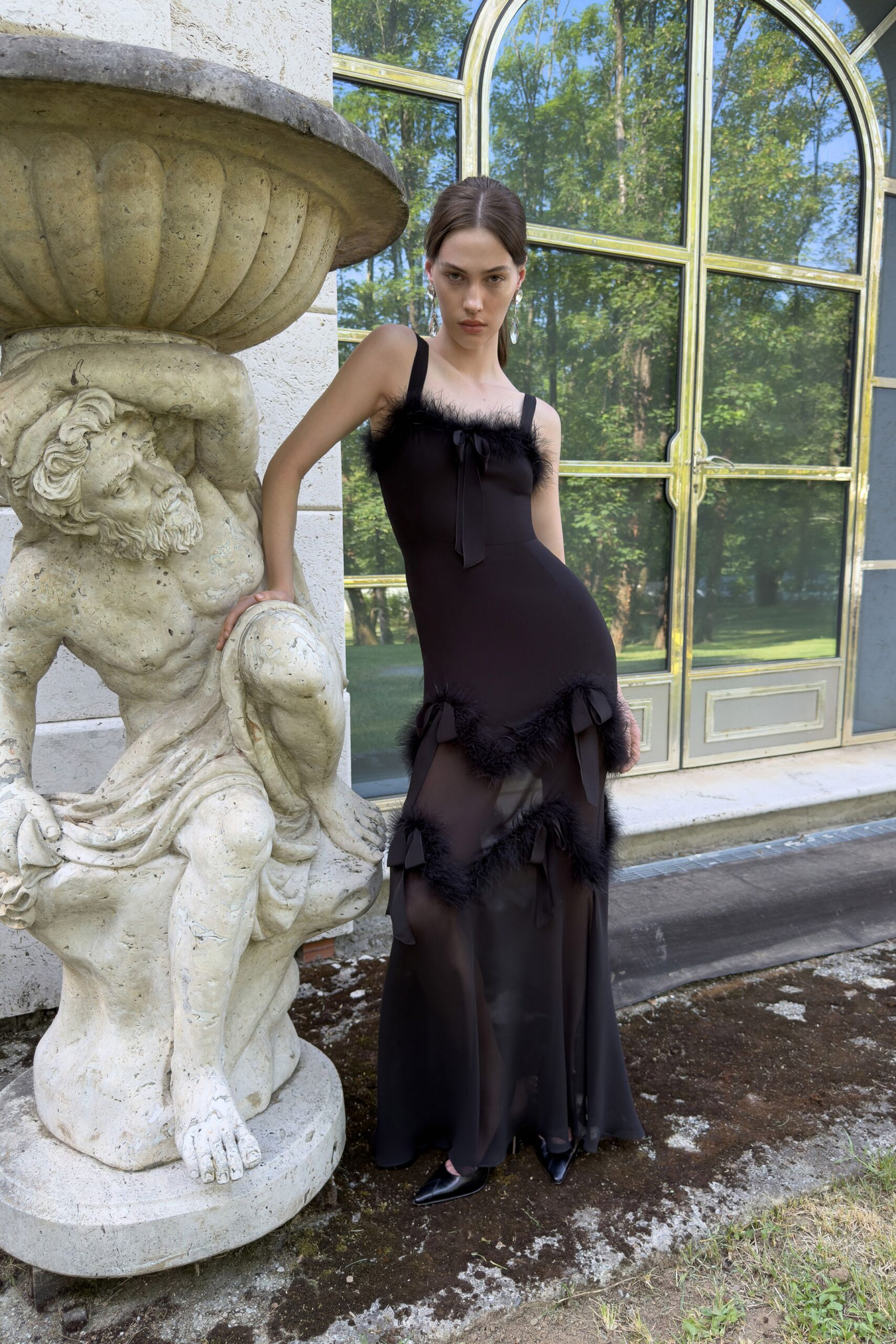 Black column gown with marabou-trimmed neckline, sheer godets tied in loose bows. Long crystal drops brushing the jaw. Black pointed pumps under the pooling hem. Crossing the orangery terrace before anyone opens the champagne.