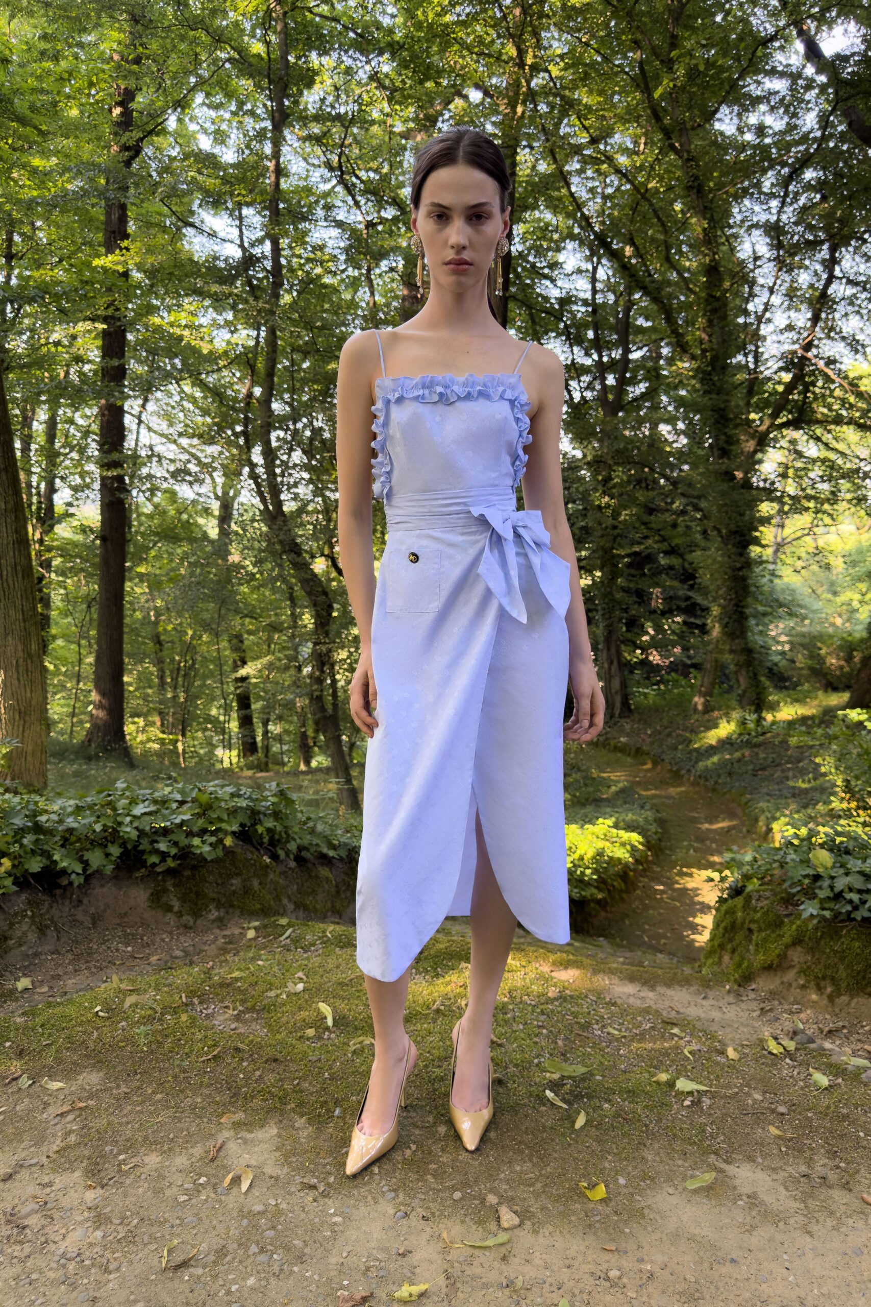 Powder-blue dress with ruffled square neckline, wrapped skirt and oversized waist bow. Crystal drop earrings brushing the jaw. Nude patent pumps with a sharp pointed toe. Walking a shaded garden path before the wedding guests find the lawn.
