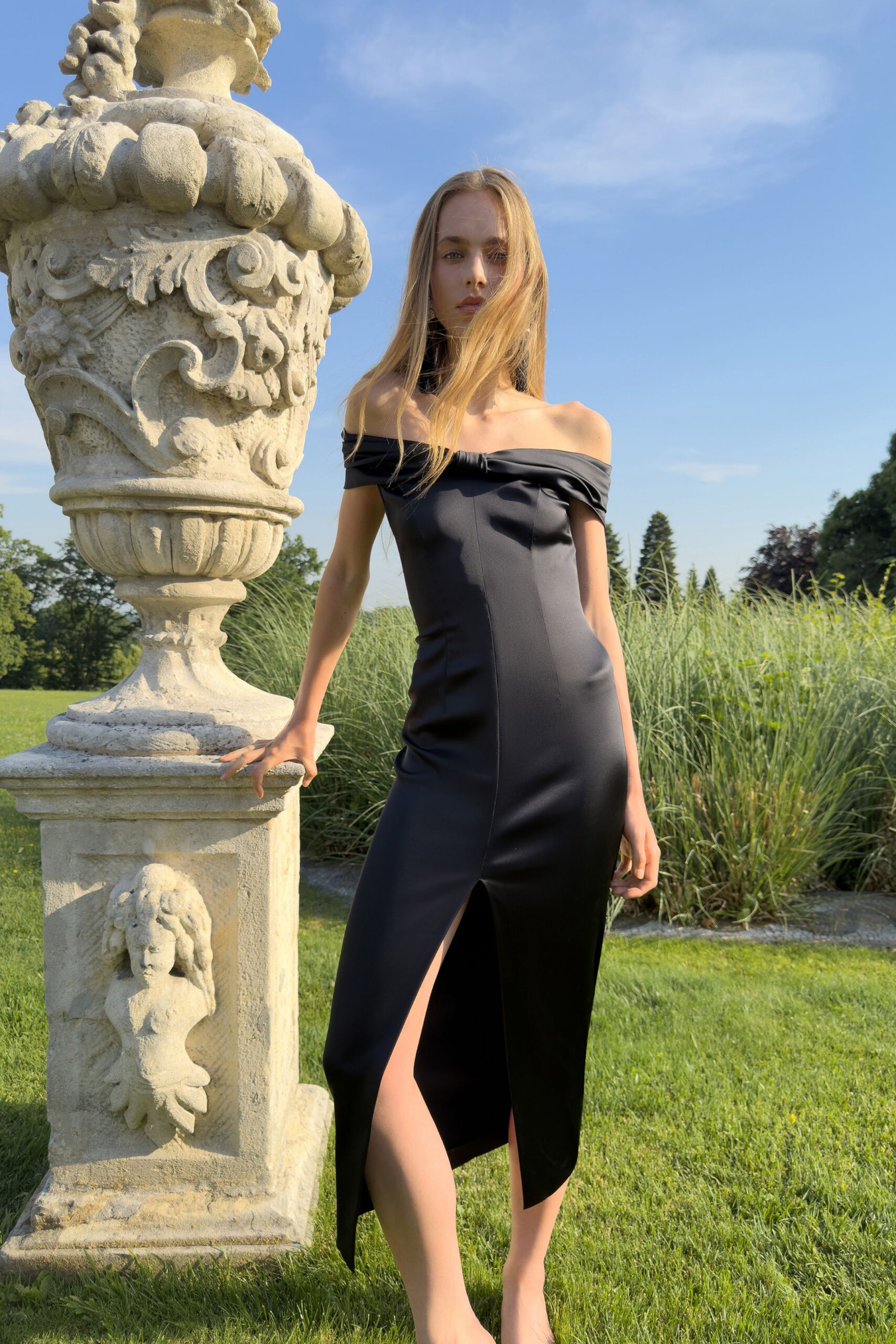 Black satin column dress with folded off-shoulder band and sharp front slit. Long black tassels brushing the neck. Crossing a formal garden before the wedding guests find the lawn.