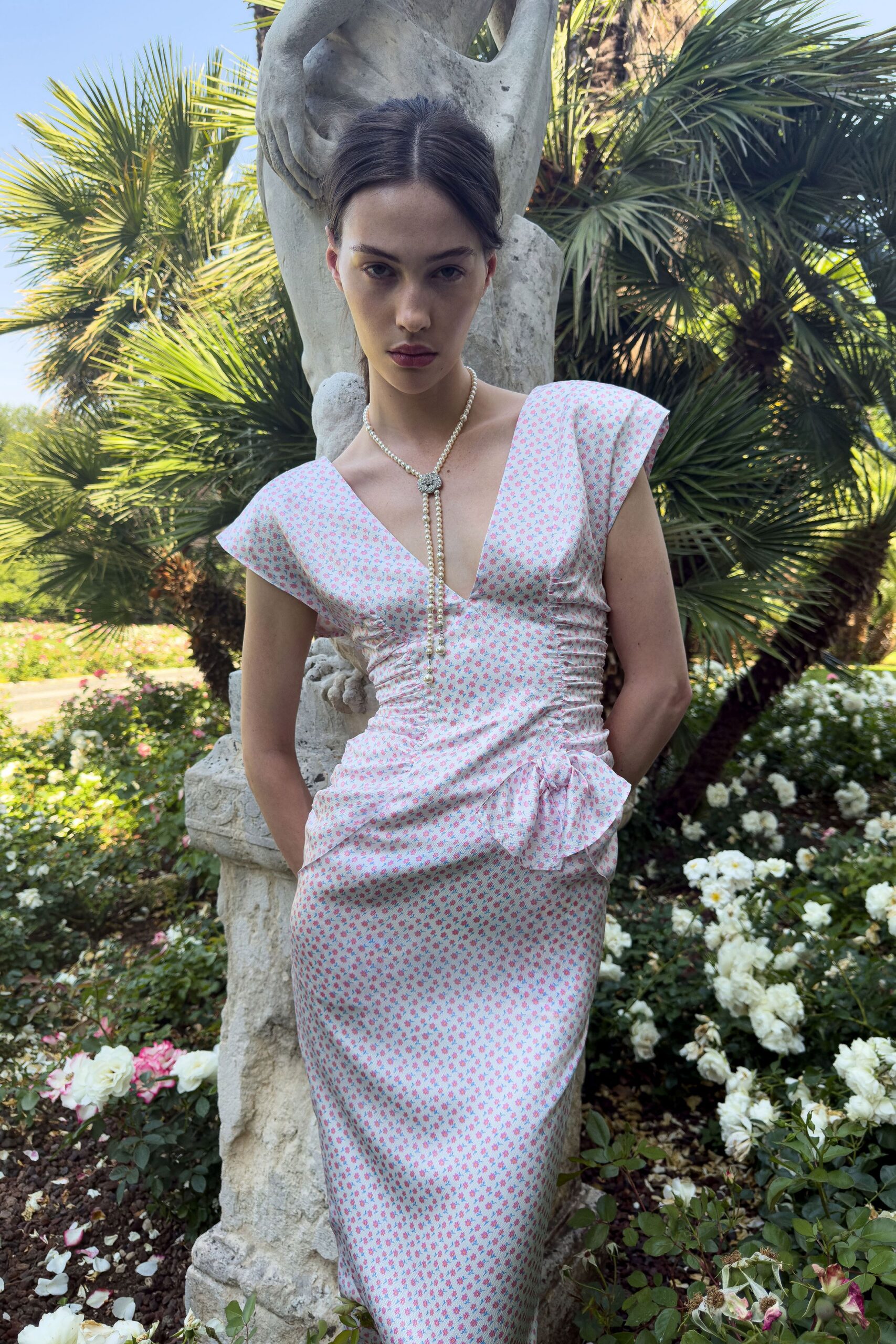 Long pearl lariat with a pavé heart clasp at the throat. Pale pink floral dress, ruched through the waist with a side bow knot. Rose garden noon, the fountain switched off for cleaning.