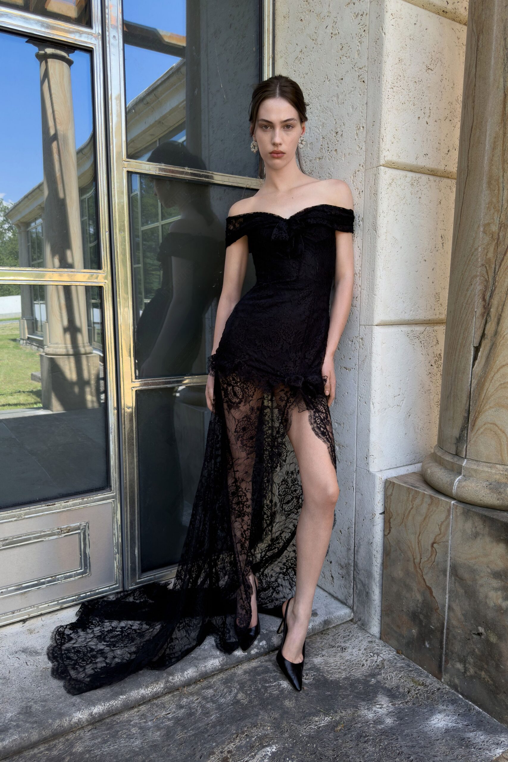 Black velvet off-shoulder gown, corseted through the waist with a sheer lace train. Clustered crystal drop earrings. Black satin pumps, needle-sharp toes. On the stone terrace before guests spill out for champagne.