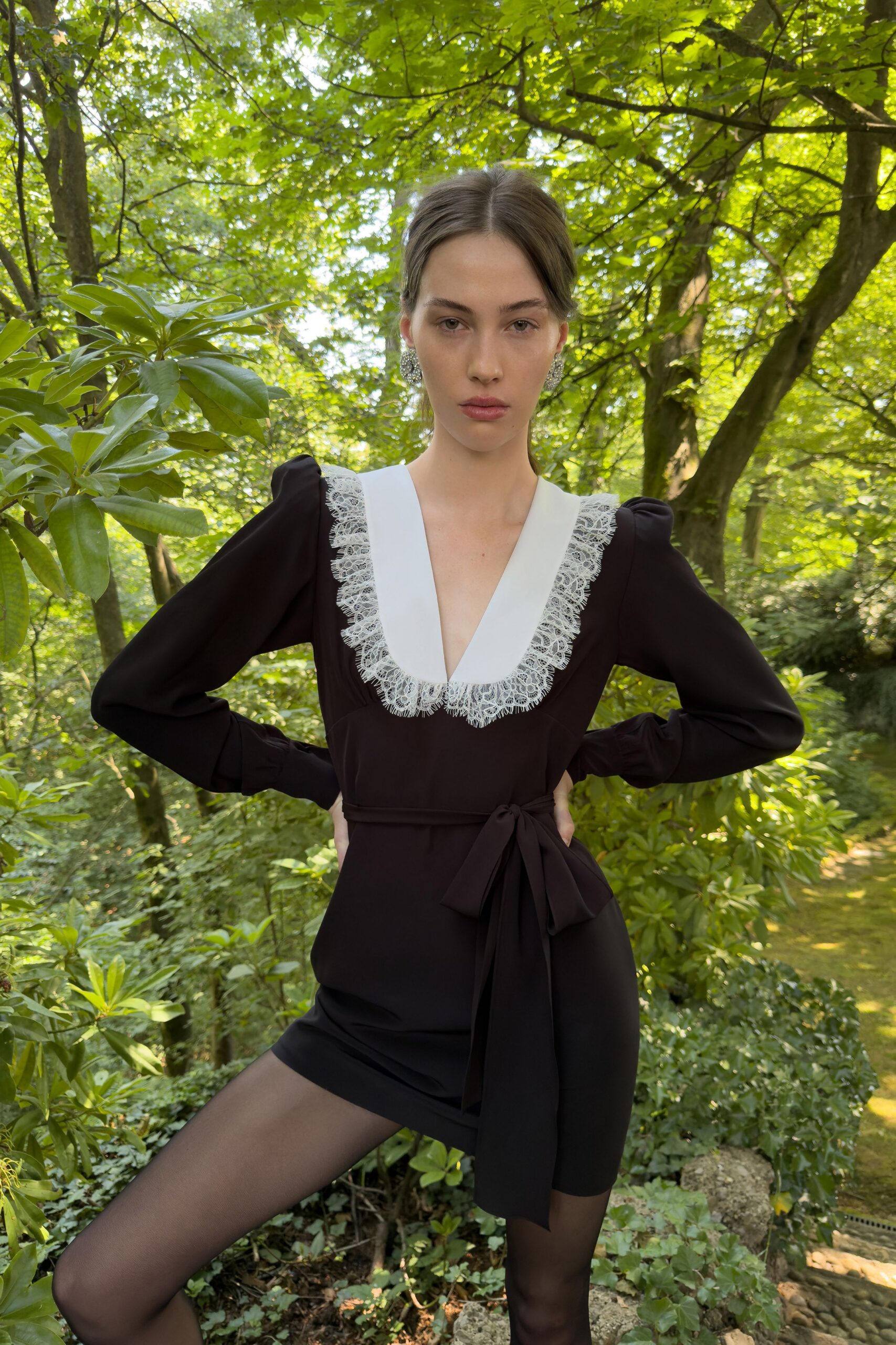 Black crepe mini dress with puffed shoulders, white bib plunging to lace. Self-fabric sash knotted low, one tail hanging past the hem. Sheer black tights. Clustered crystal earrings. Walking the mossy path behind a country house after everyone else went in.