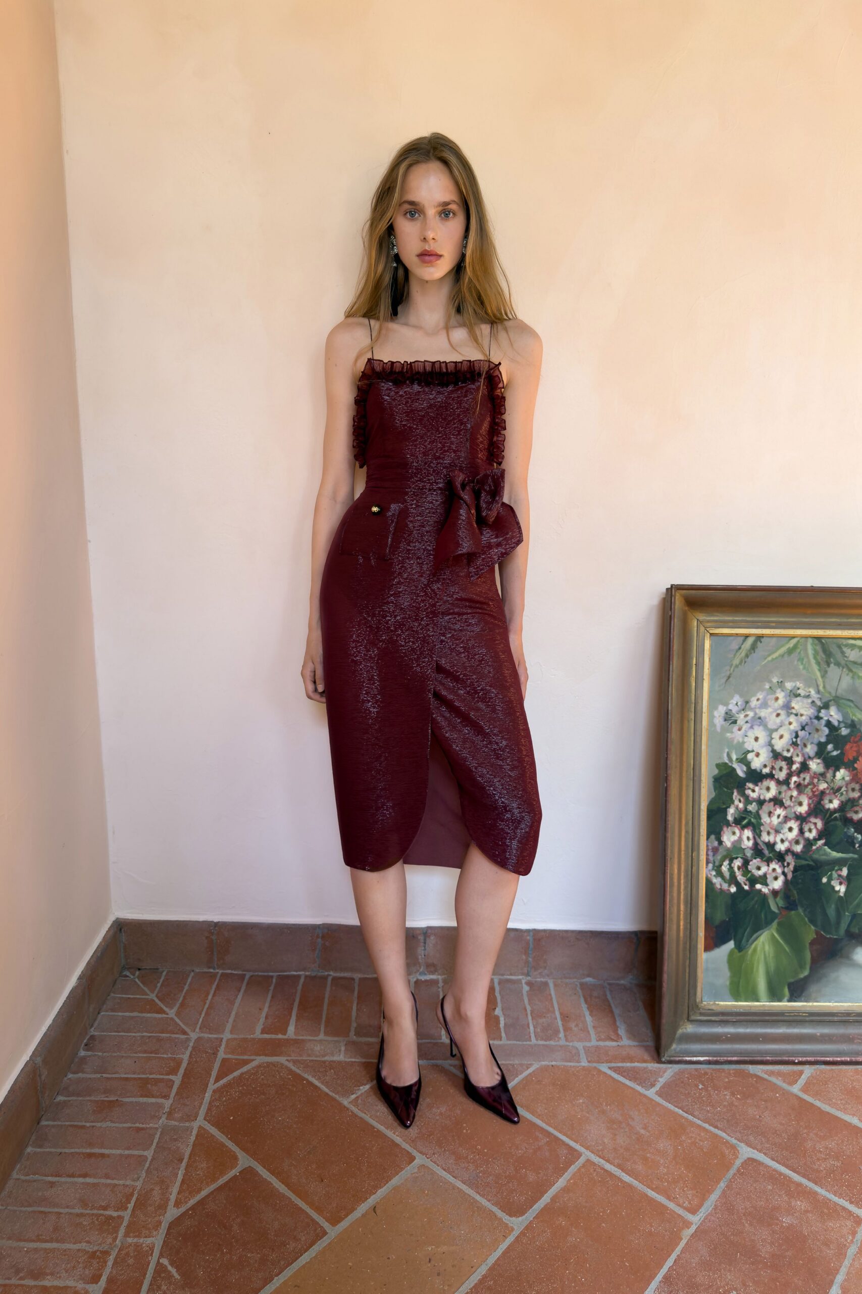 Burgundy shimmer-knit slip dress, organza ruffle neckline and a bow at the hip. Pointed oxblood satin pumps. Standing in a tiled villa hallway before the dinner guests spill onto the terrace.