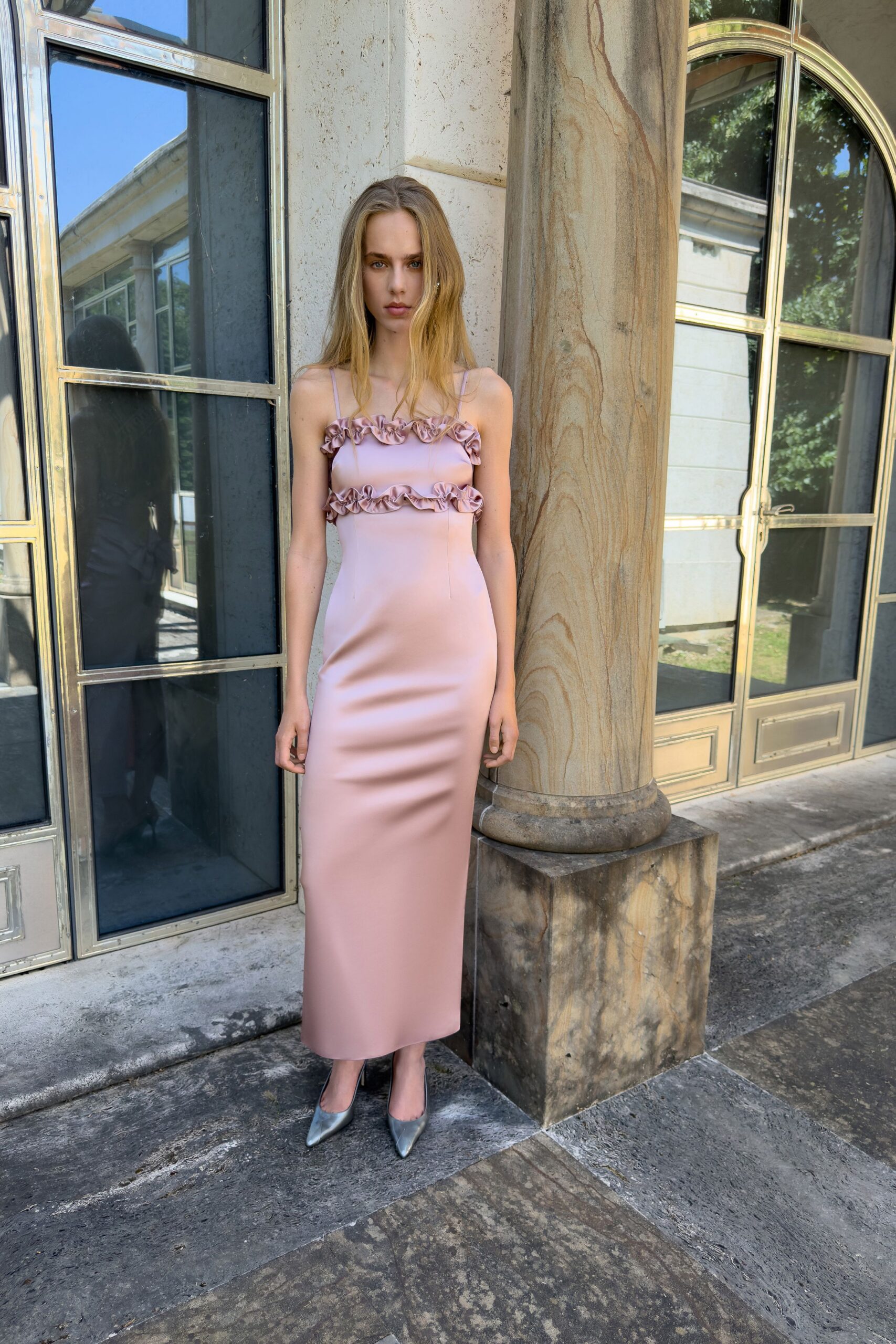 Blush satin slip dress, two ruffled bands rippling across the straight neckline and bust. Silver pointed pumps with mirror-shine leather. Crossing a stone courtyard before the wedding guests have found their seats.