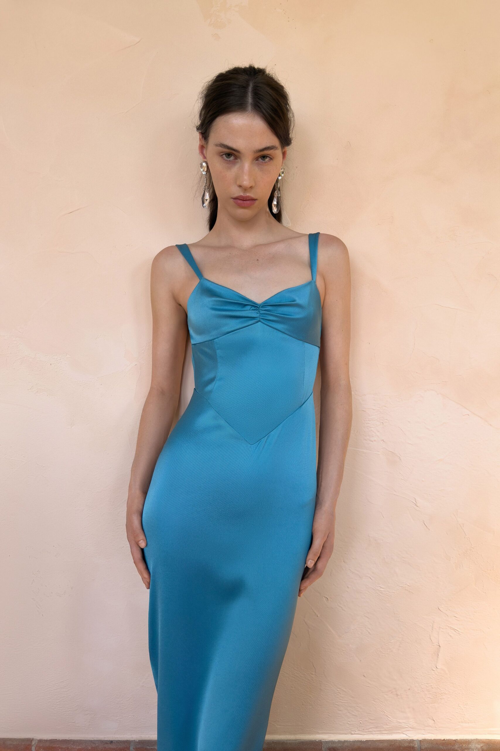 Faceted drop earrings catching light beside the jaw. Turquoise satin slip dress, ruched sweetheart bust and pointed midriff panel. Terracotta corridor at noon, on the way to somebody else's wedding.