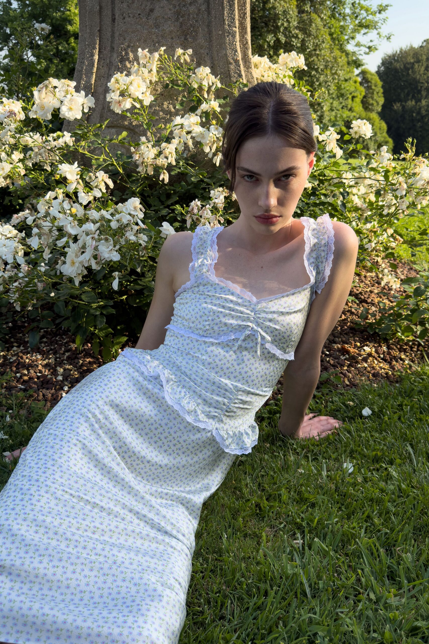 White cotton slip dress scattered with tiny lilac sprigs and lace-edged ruffles. Wide shoulder straps trimmed in scalloped white lace. Ruched sweetheart bodice cinched by a narrow tie at center. Dropped waist peplum flicking out in a soft ruffle. Rose garden grass still warm after the wedding guests have drifted inside.