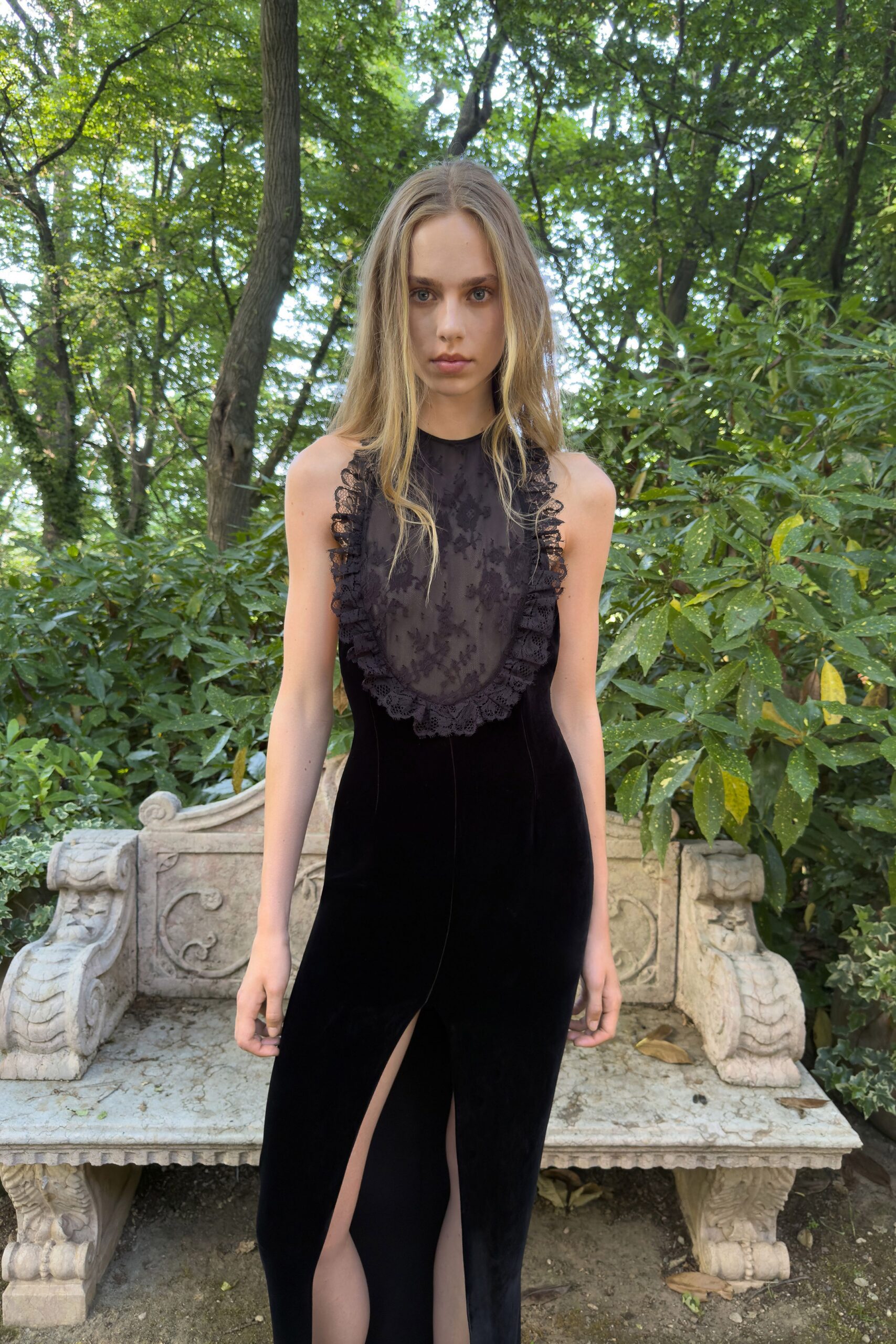 Black velvet column gown with a sheer flocked bib framed in ruffled lace. Front slit opening in two narrow panels from mid-thigh. Sitting on a stone garden bench after the wedding guests have wandered off.