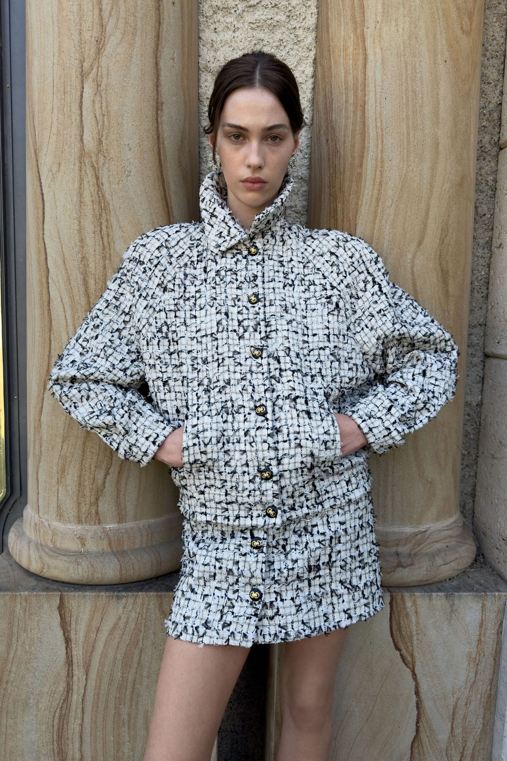 Crystal cluster drop earrings. White-and-black bouclé jacket with a raised funnel collar and crest buttons. Matching bouclé mini, frayed hem brushing high on the thigh. Stone steps outside a civic building, noon sun catching in the grain.