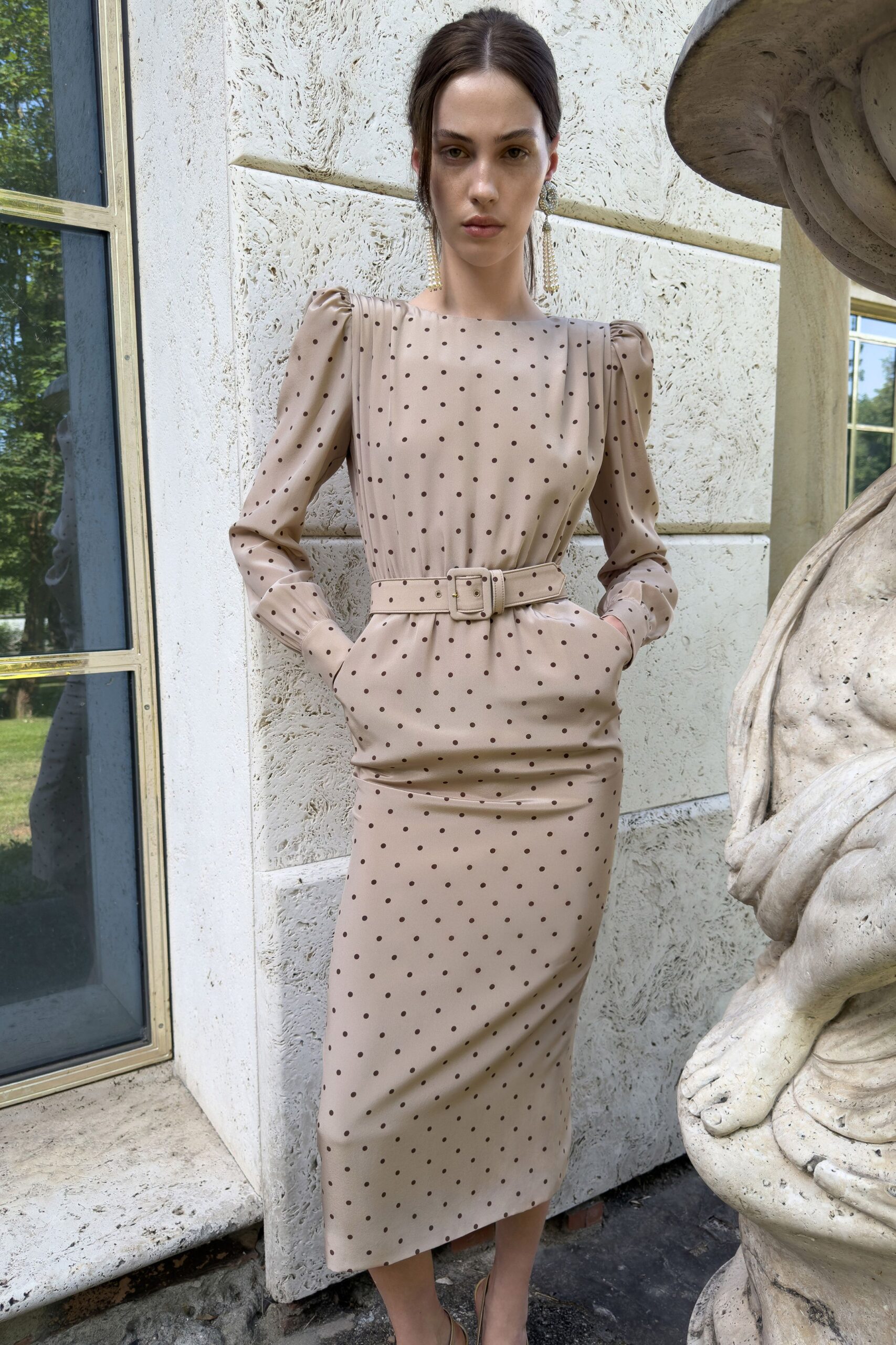 Crystal cluster drops ending in thin pearl tassels. Blush crepe dress scattered with brown pin dots, puffed shoulders and ruched hips. Self-covered belt with a square buckle cinching the waist. Barely there ankle-strap sandals. Garden wedding reception, standing beside the fountain before speeches start.