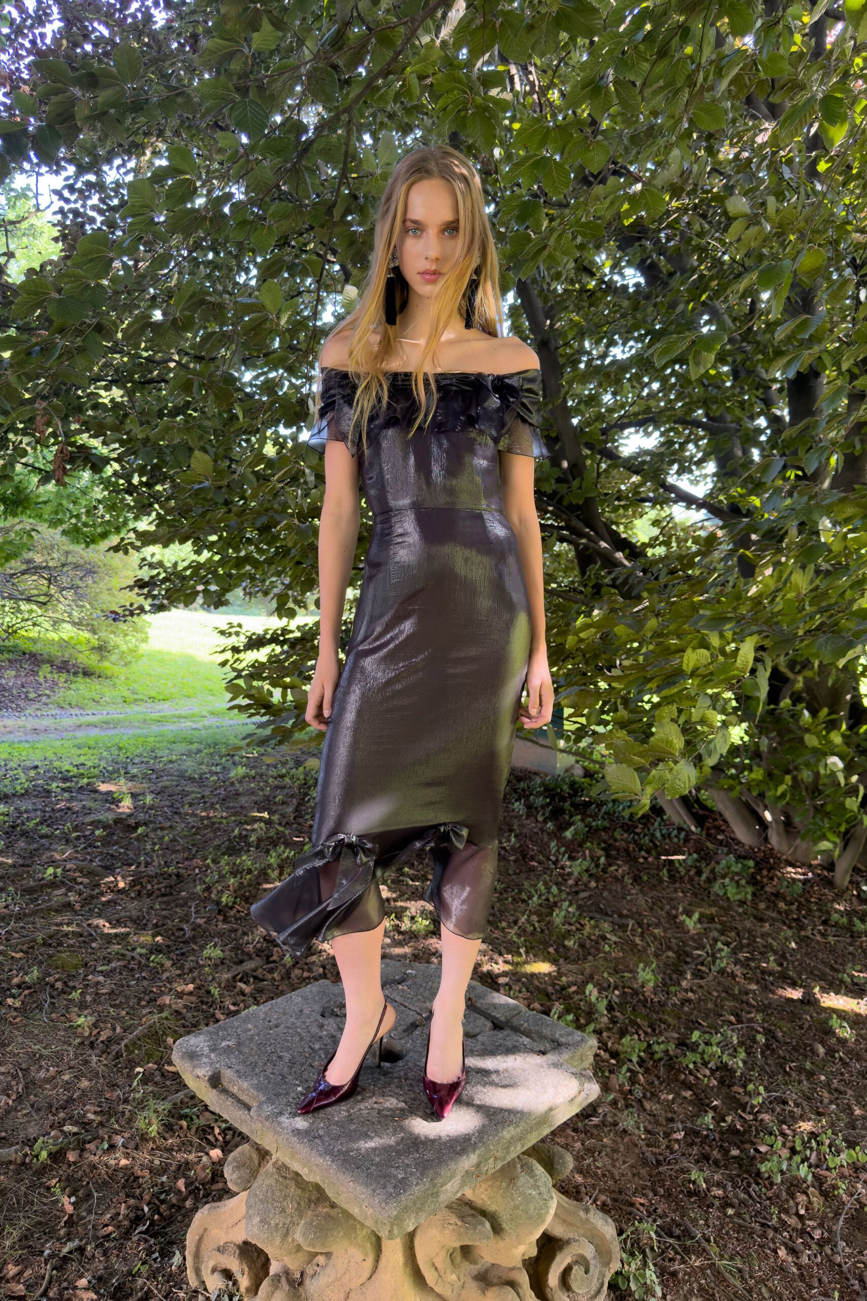 Long black tassel earrings with clustered crystal tops. Off-shoulder black lamé dress, ruffled neckline and one bow snagging the hem. Oxblood patent slingback pumps with knife-point toes. Standing on a stone garden table after the wedding strayed into dusk.